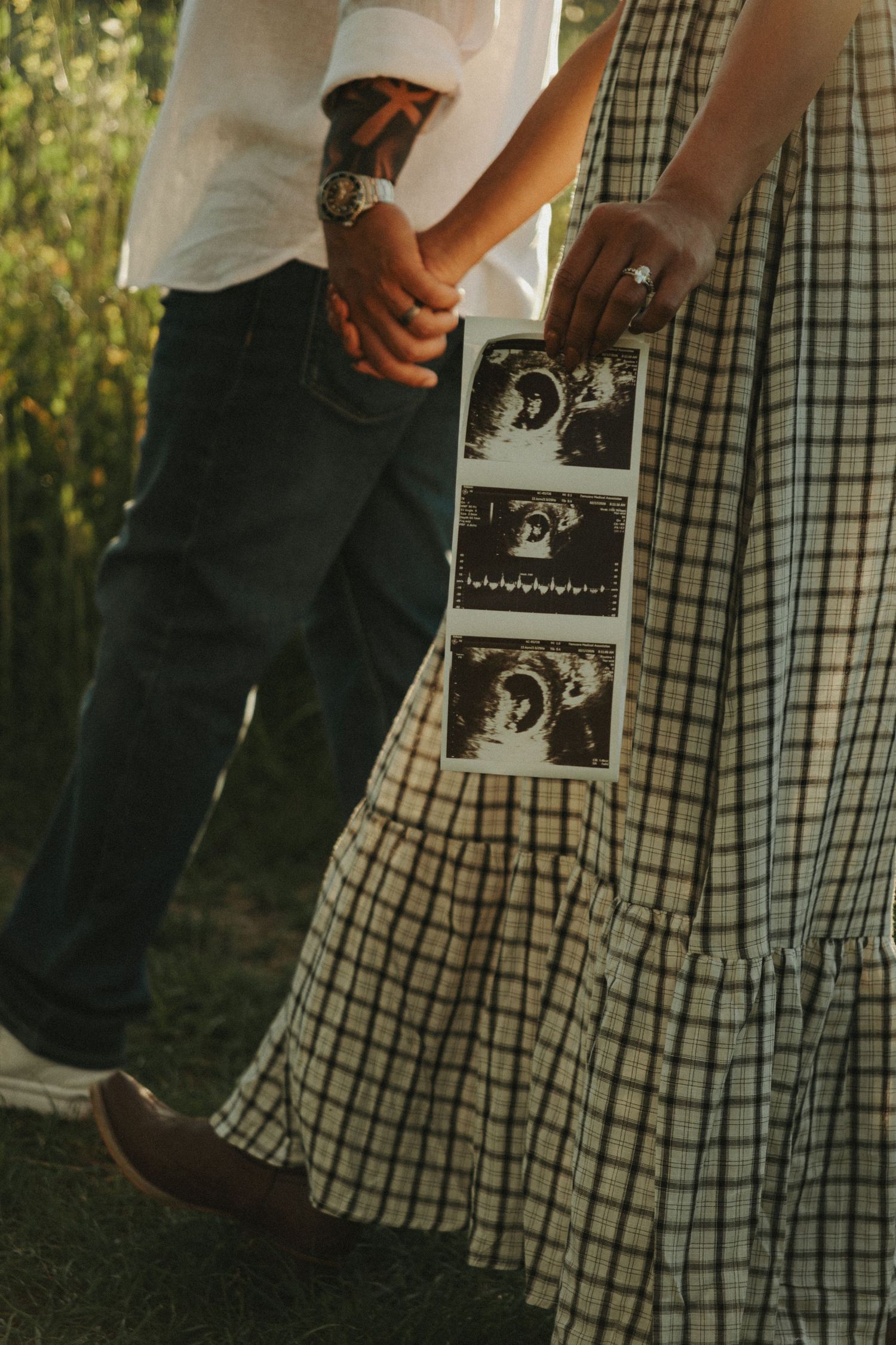 Couple holding hands outdoors displaying ultrasound photos, pregnancy announcement with checkered dress and tattooed partner.