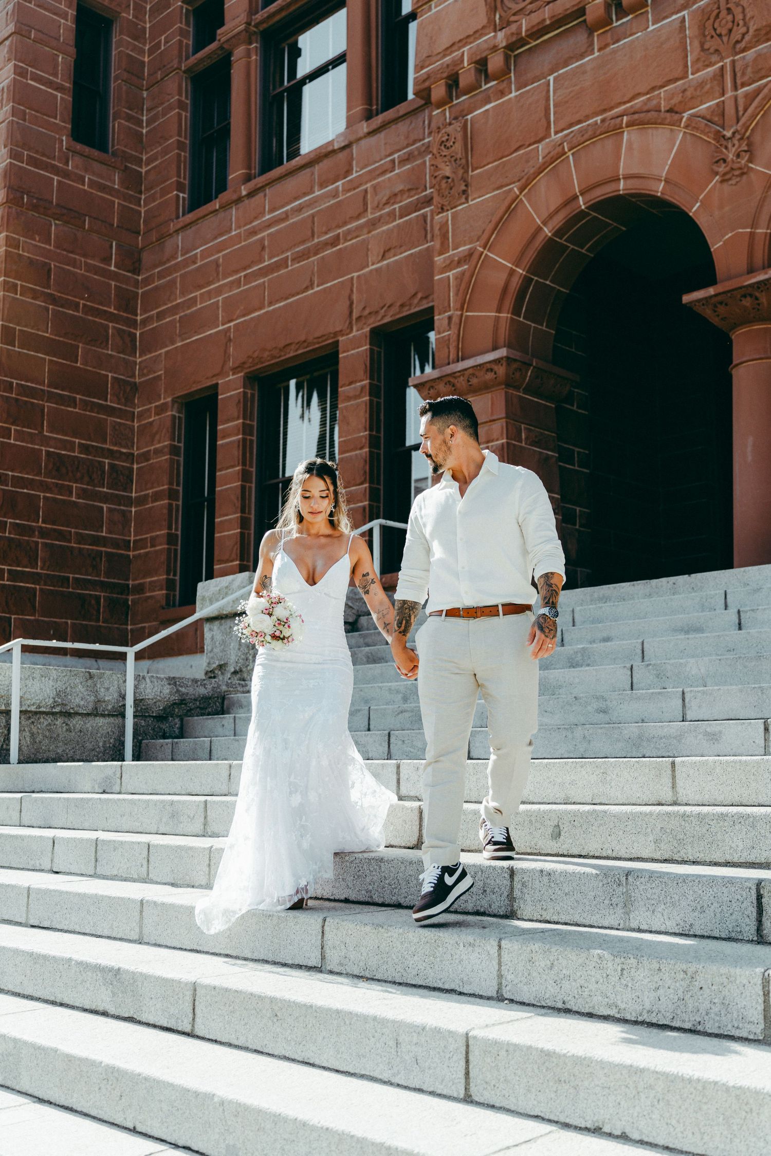 A couple in white wedding attire sits on stone steps in front of a brick building.