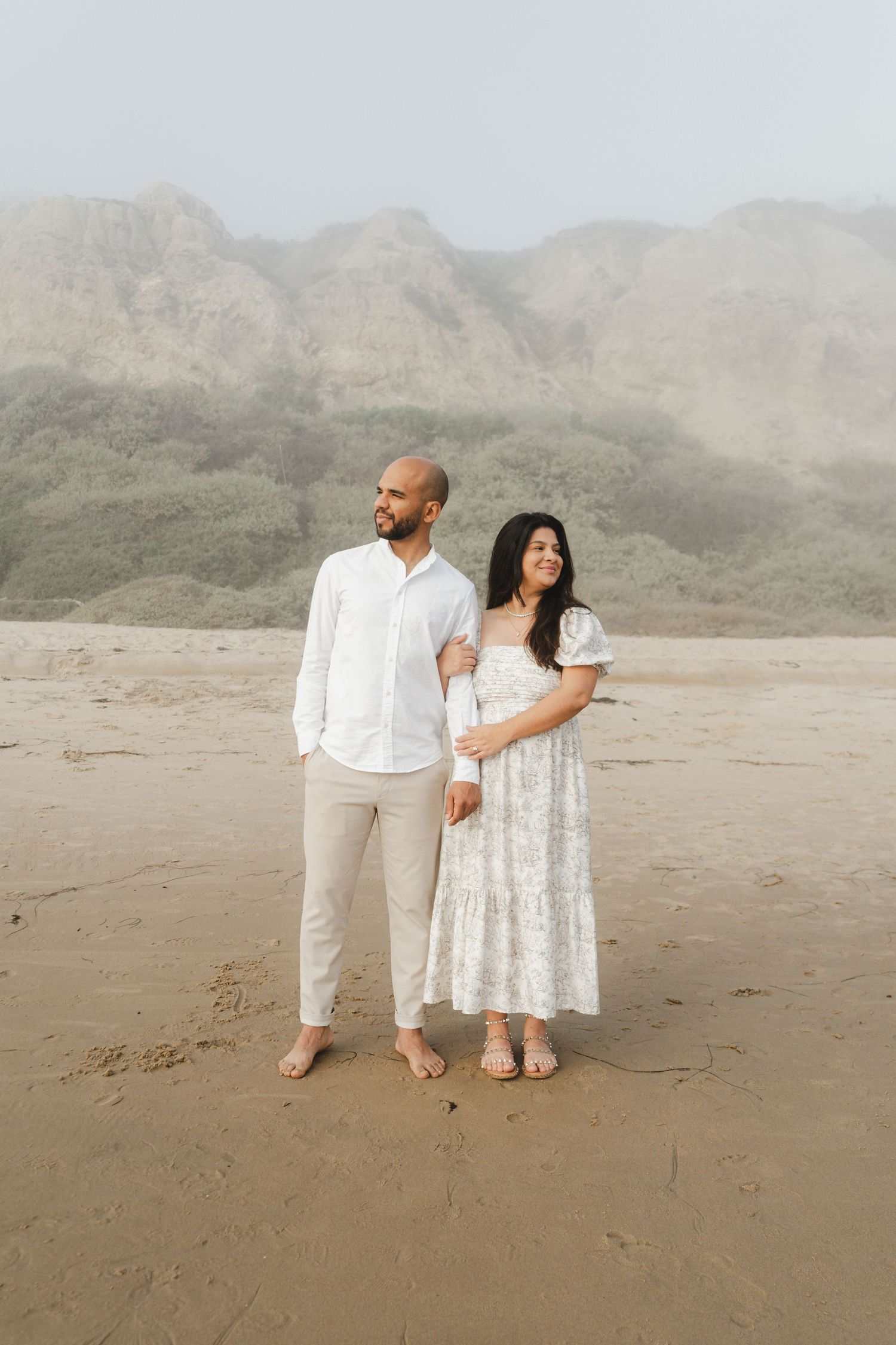 A couple in coordinated white outfits stands against a backdrop of desert mountains.
