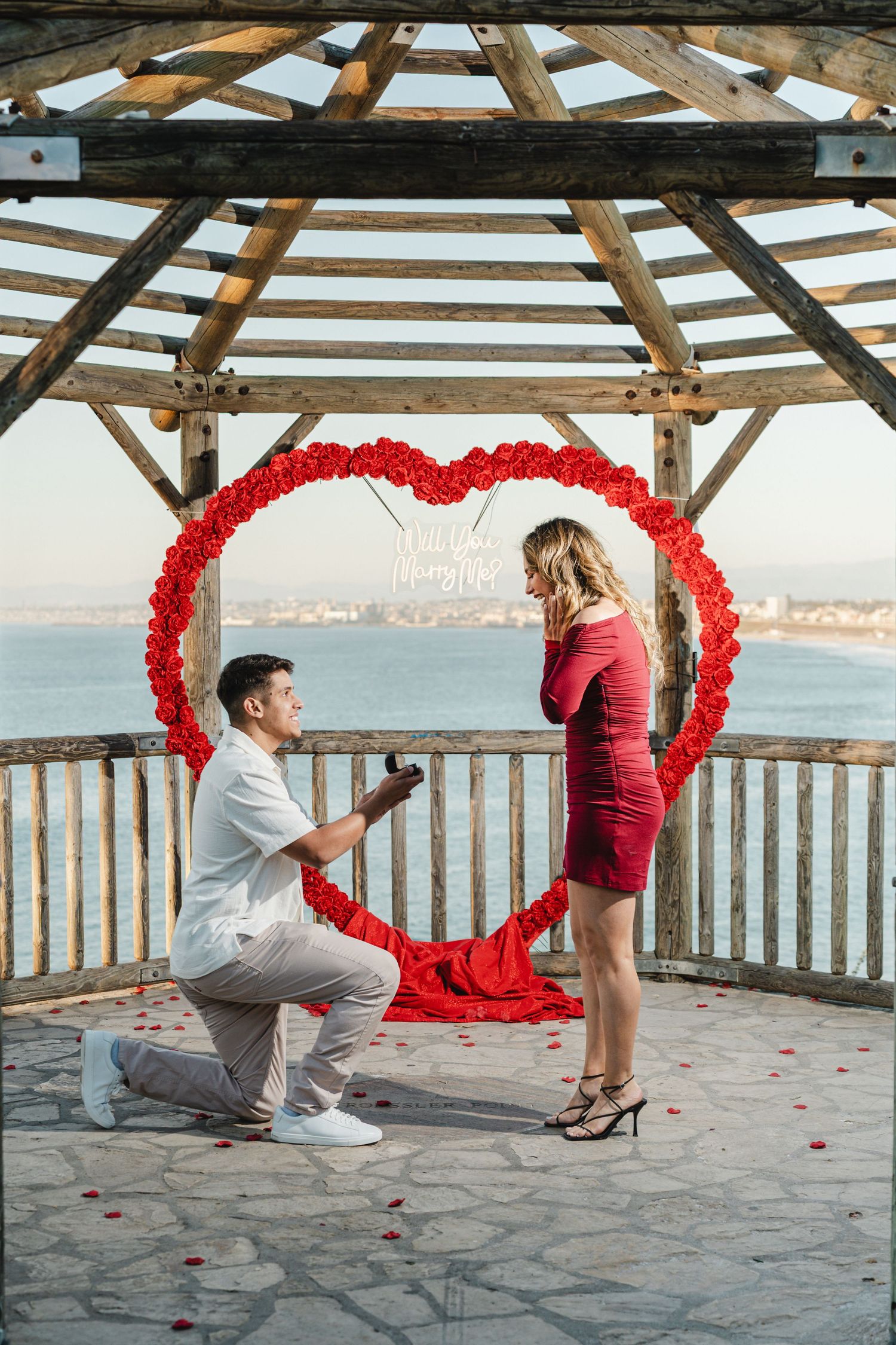Marriage proposal scene with red heart decoration and rose petals on oceanfront wooden deck.