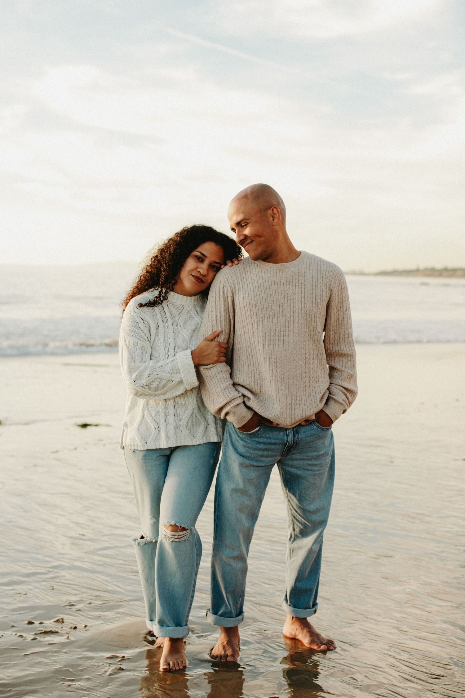 Couple wearing matching sweaters and jeans standing together at the beach during sunset.