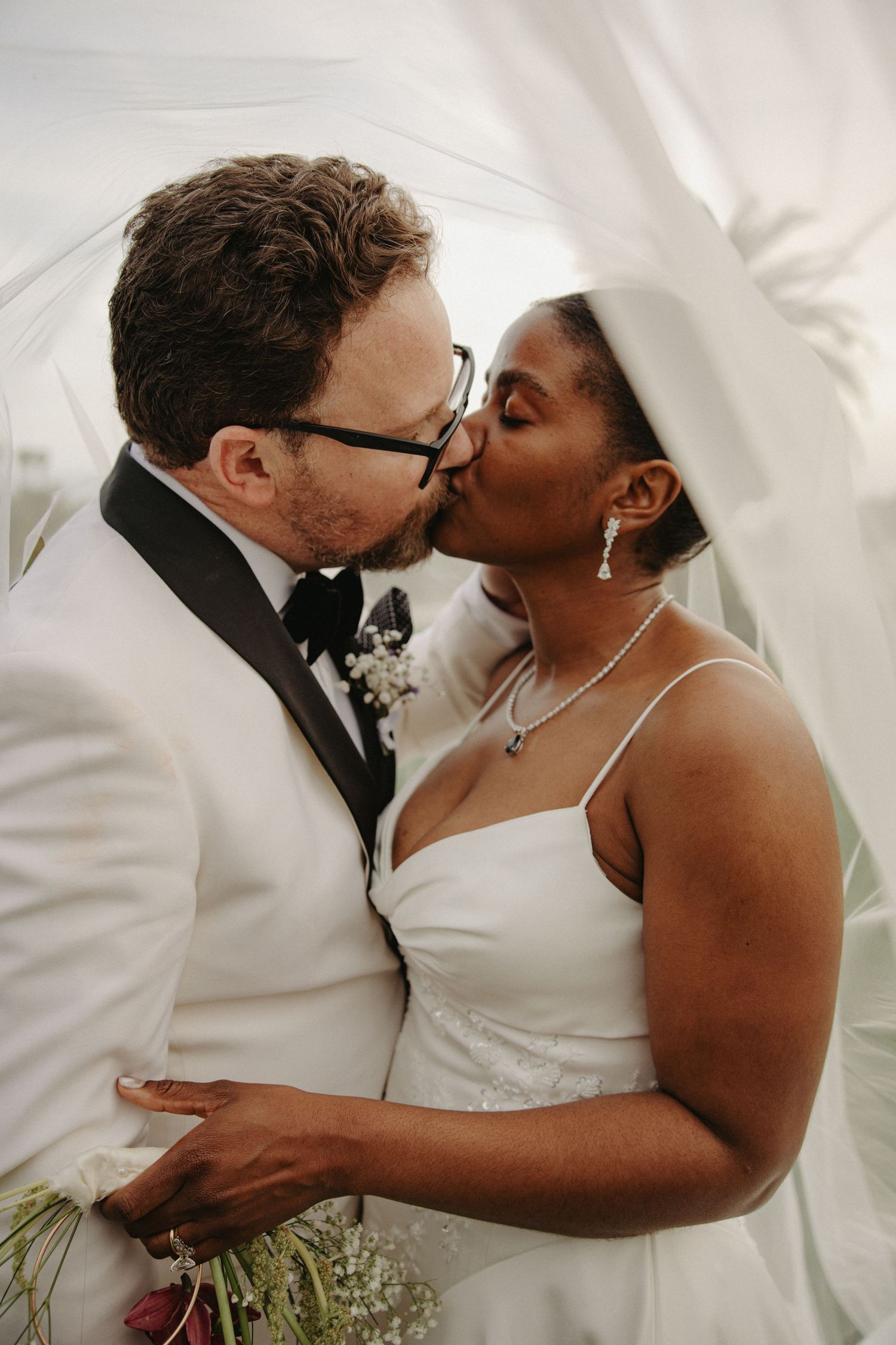 Interracial couple sharing a romantic kiss under a flowing white veil on their wedding day.