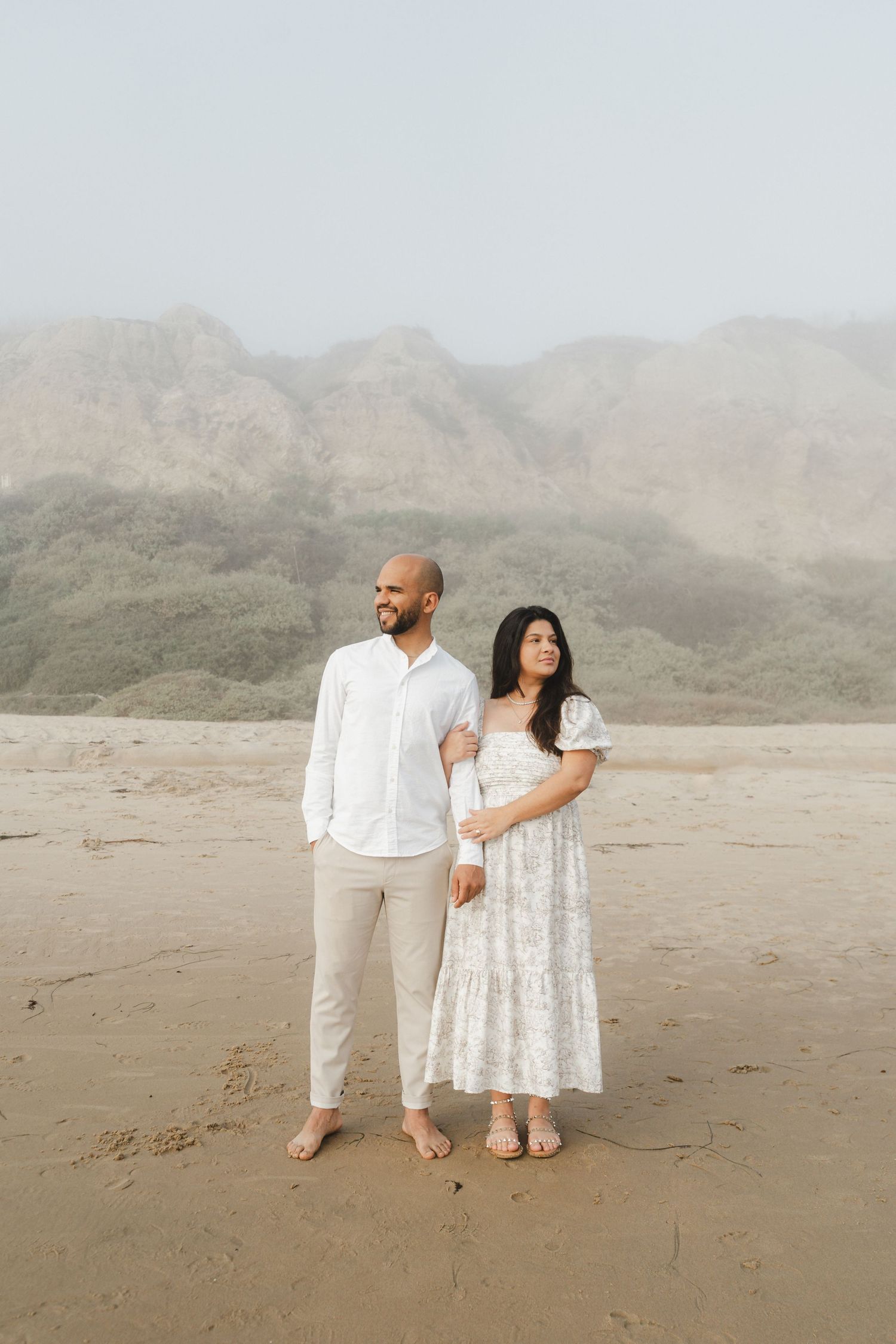 A couple in white outfits stands together on a misty beach with mountains in the background.