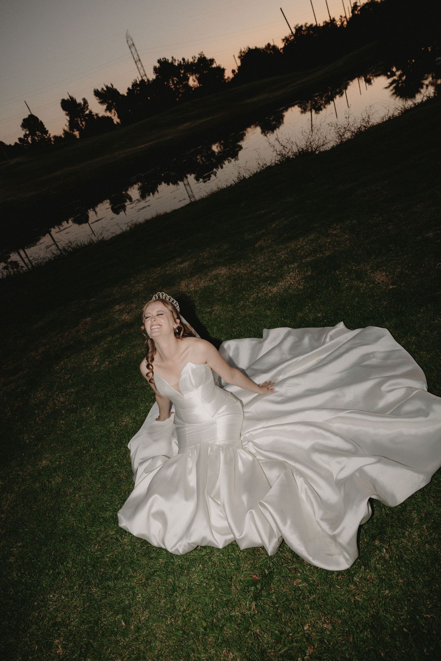 A bride in a flowing white satin gown lies on grass at dusk with a city skyline reflected in water behind her.
