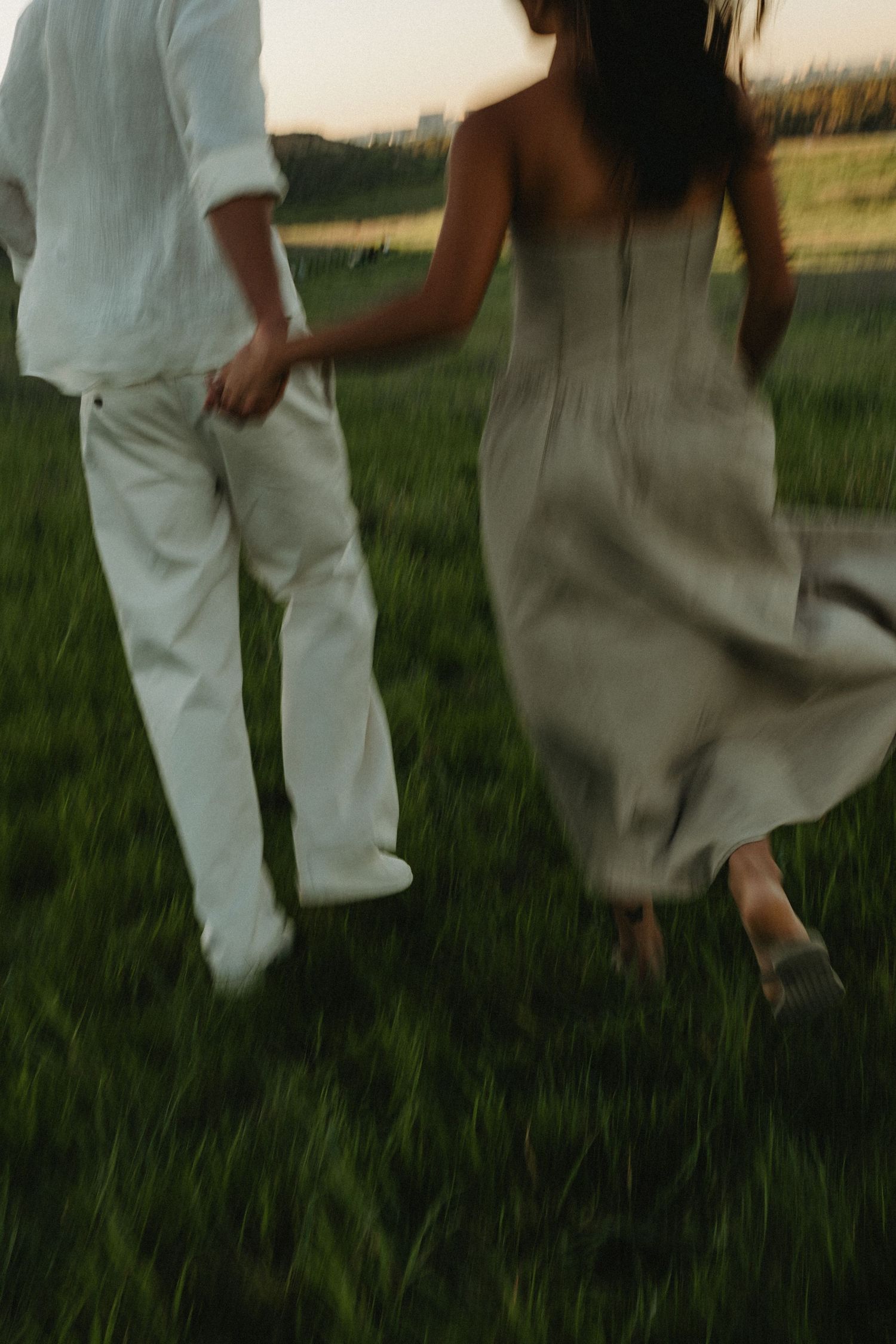 Two people walk playfully through tall grass at sunset wearing contrasting white and beige outfits.