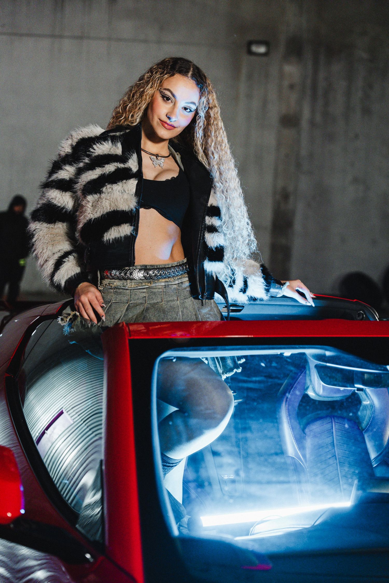 Model in striped fur jacket poses on red sports car with dramatic lighting.