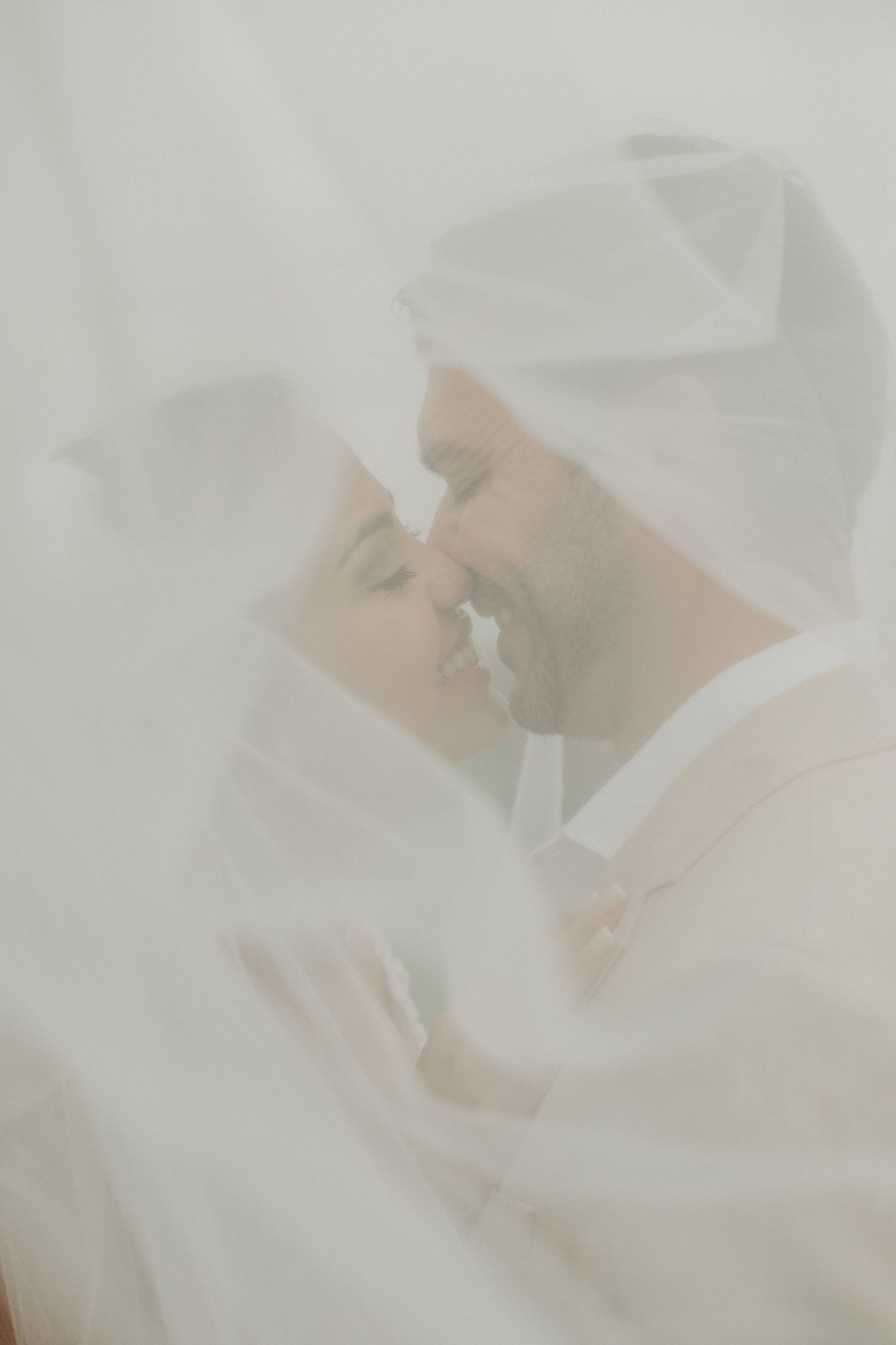A bride and groom share an intimate almost-kiss beneath a soft white wedding veil in a dreamy, ethereal portrait.