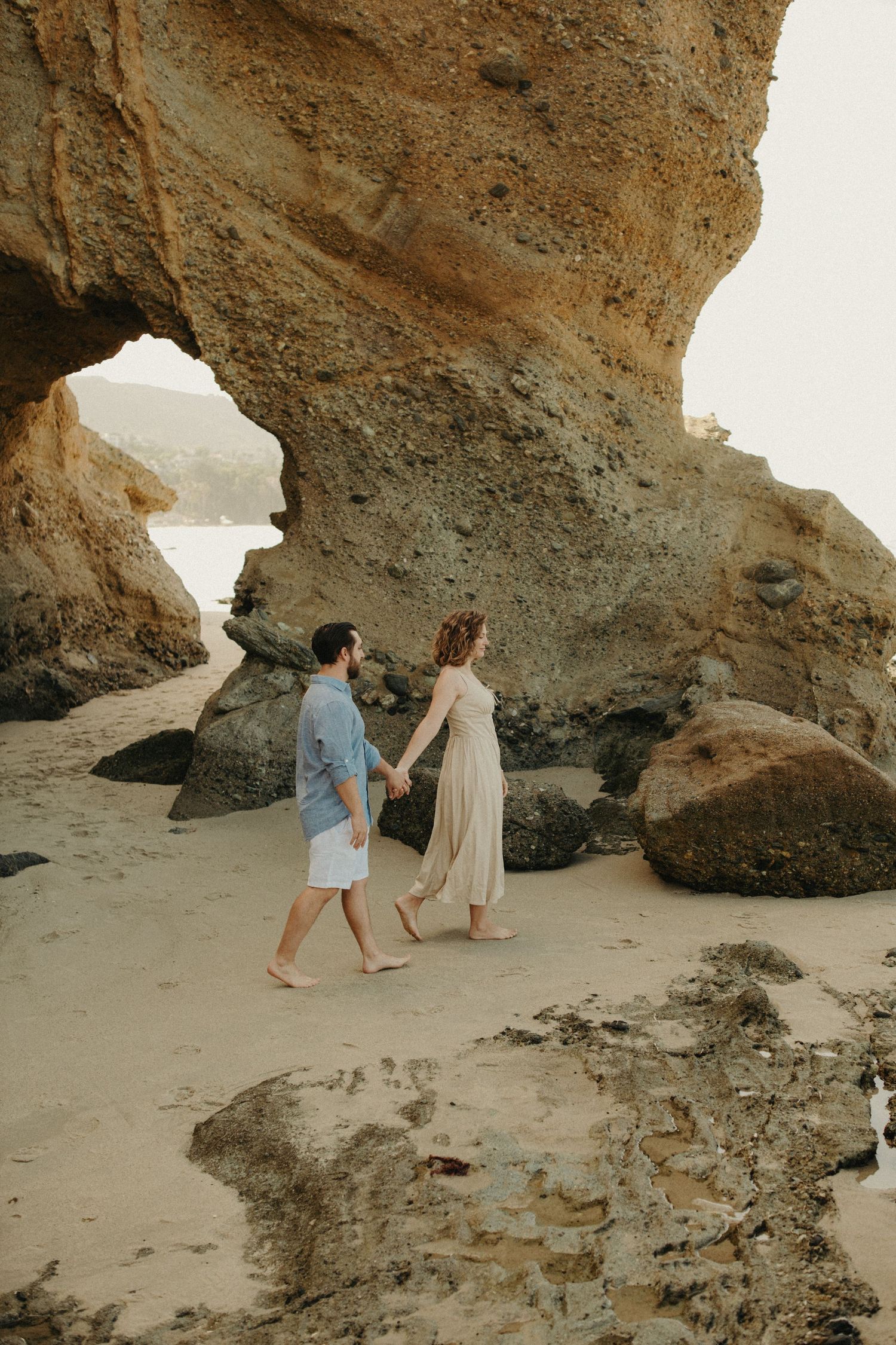 Couple holding hands walking barefoot on sandy beach beside dramatic rock formations and sea cave.