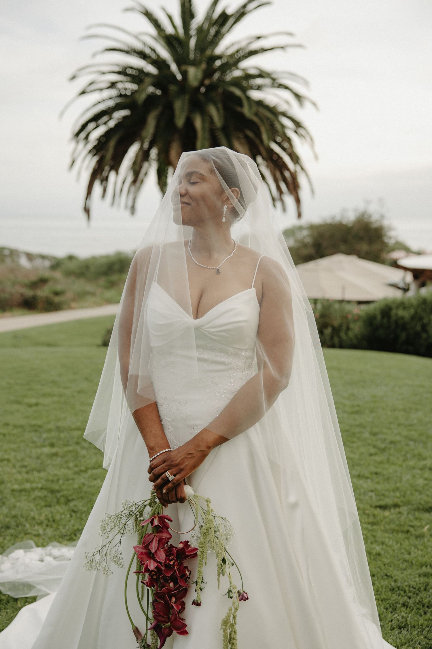 Bride in white gown and veil holds red floral bouquet outdoors, smiling with eyes closed near a palm tree.