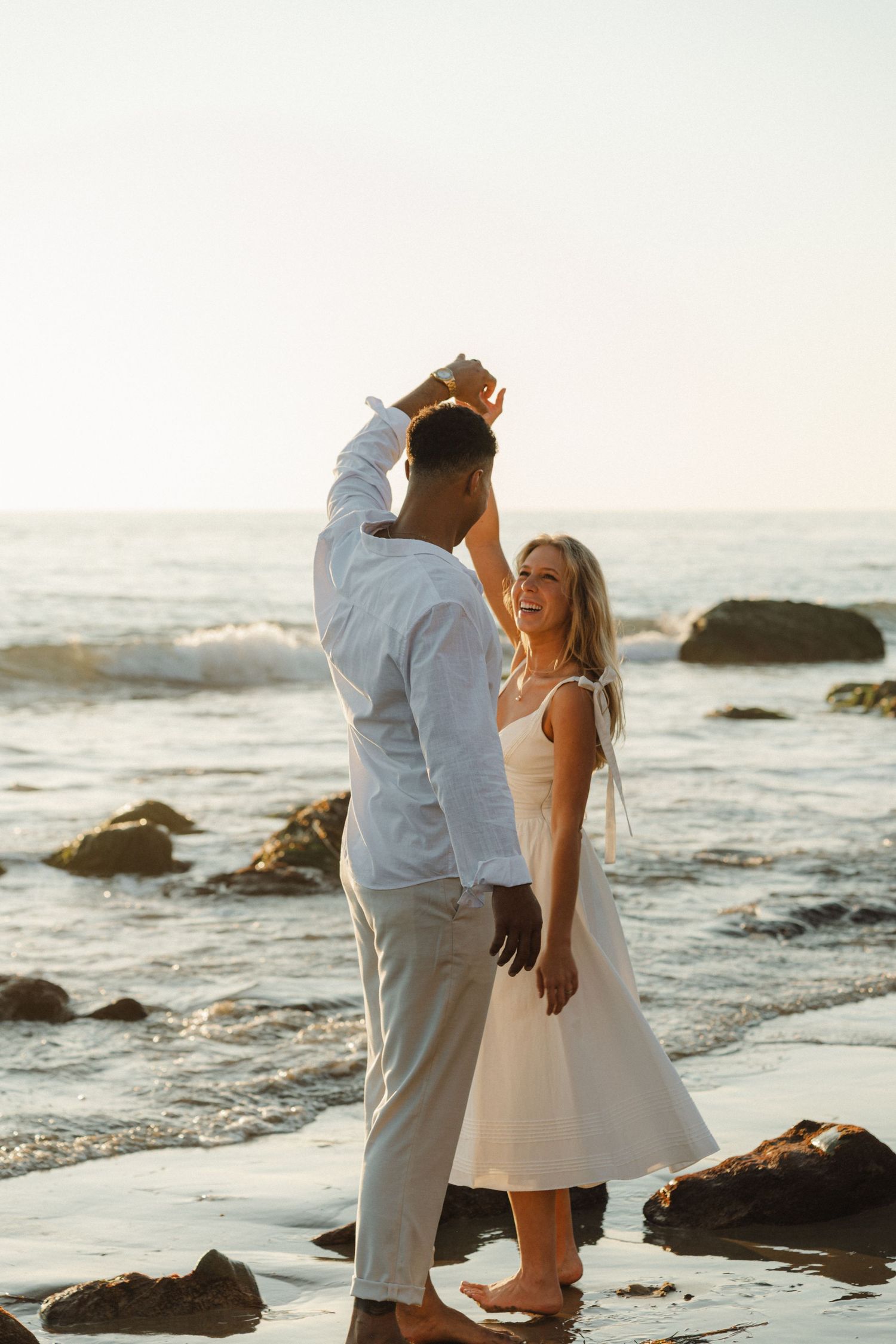 A couple shares a romantic moment on a rocky beach at sunset with waves crashing in the background.