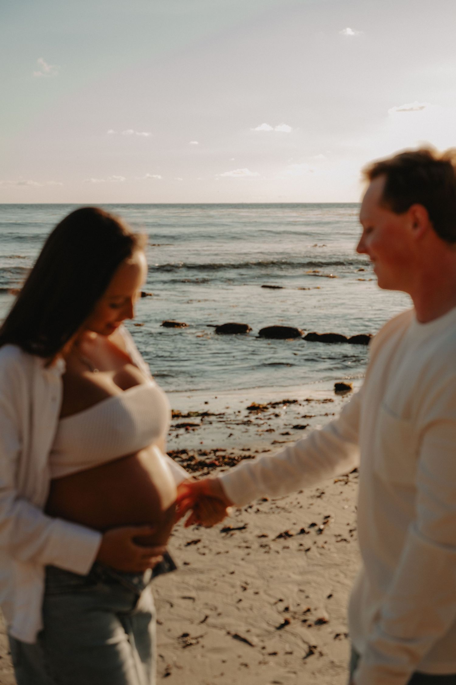 A couple embraces and dances together on a scenic beach at sunset with waves and rocks visible in the background.