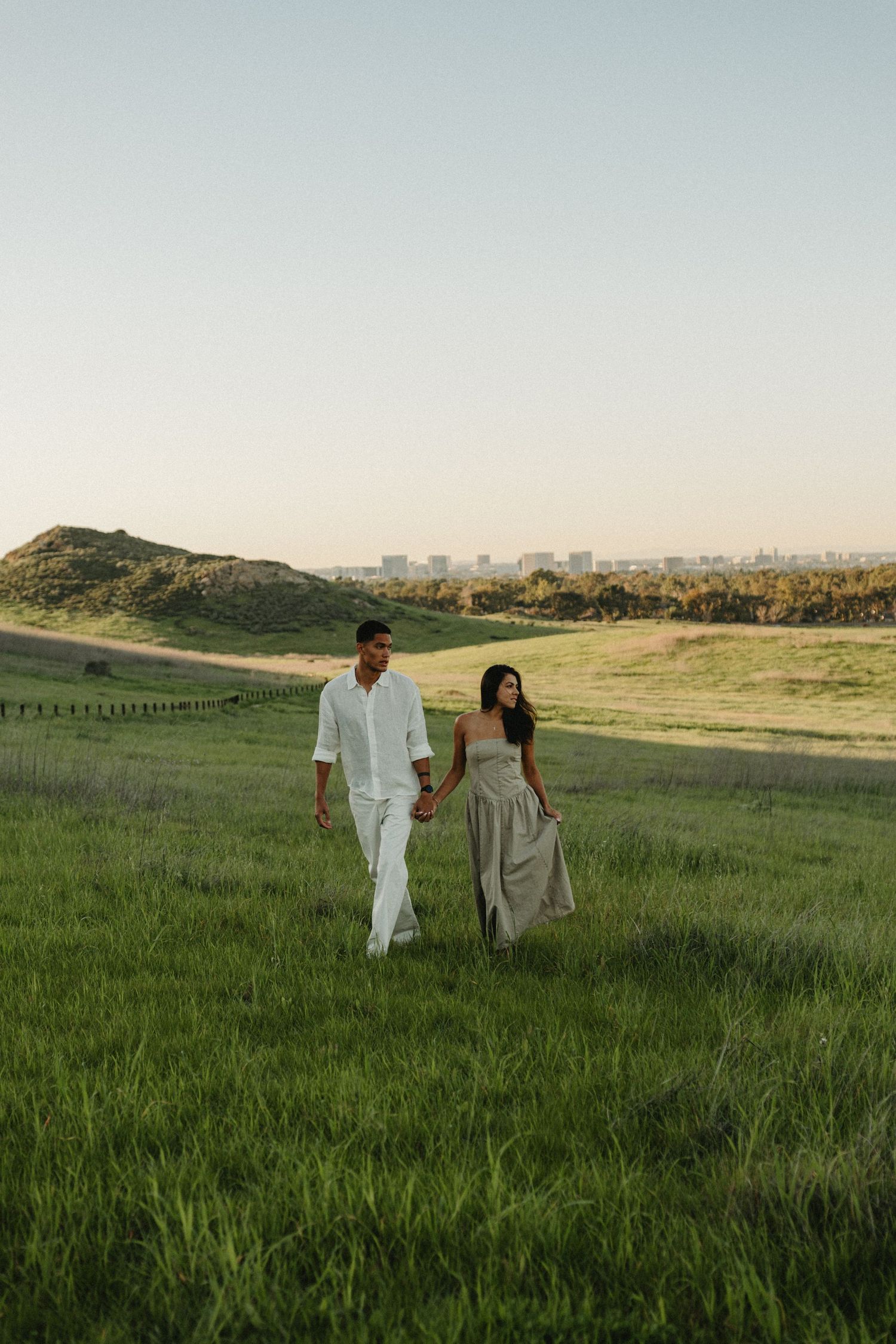 A couple walks hand in hand through a scenic meadow with a city skyline visible in the distance.
