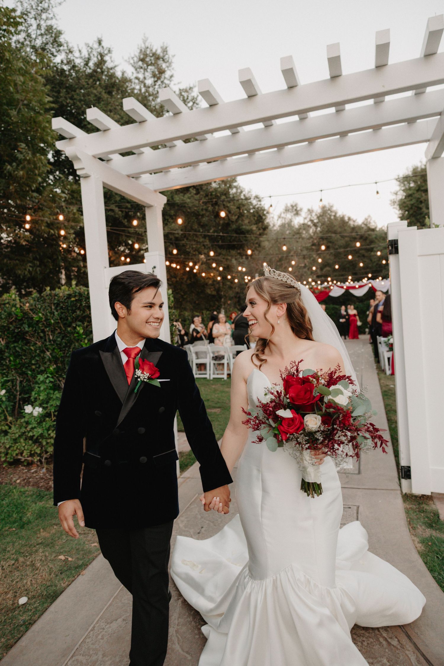 A wedding couple shares joyful moments under a white pergola decorated with string lights and red floral arrangements.