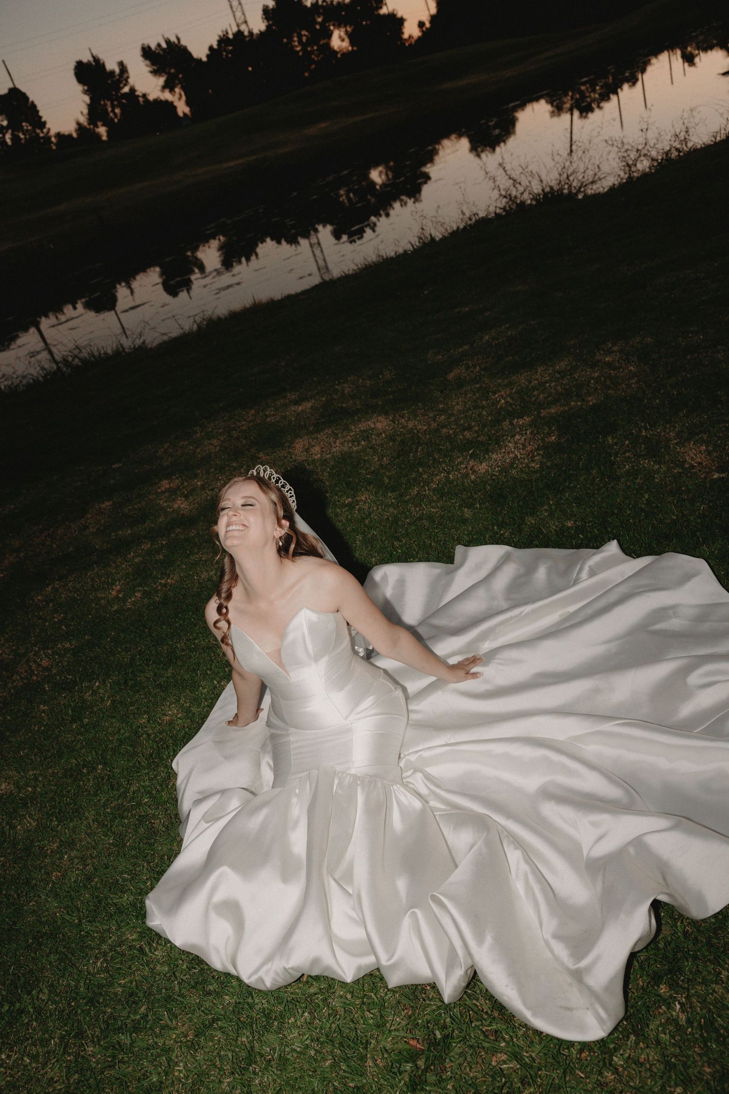 A flowing white satin wedding dress spreads across dark grass at dusk, creating a dramatic contrast.