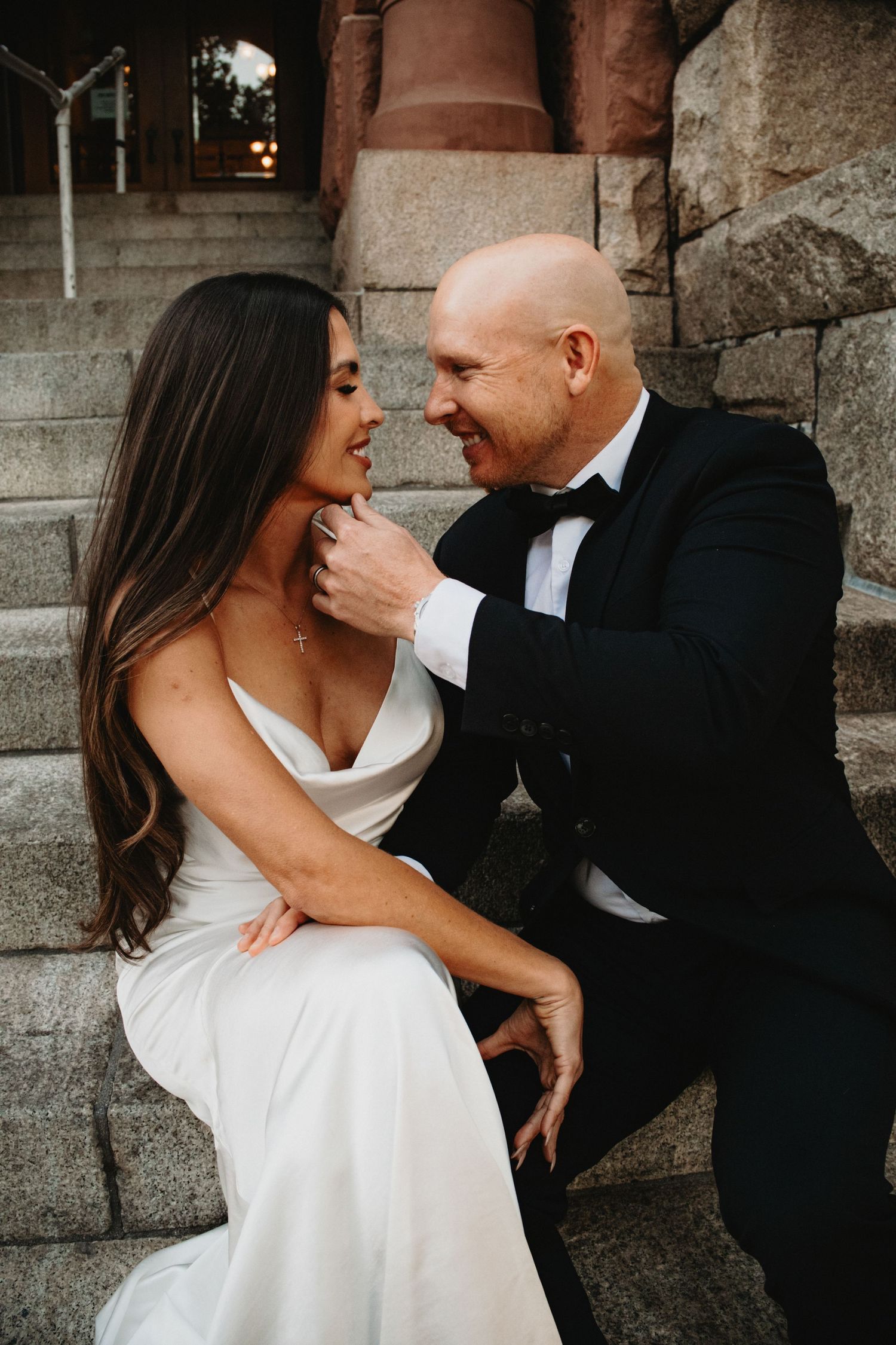 A joyful couple shares an intimate moment on stone steps, the bride in white and groom in classic black tuxedo.