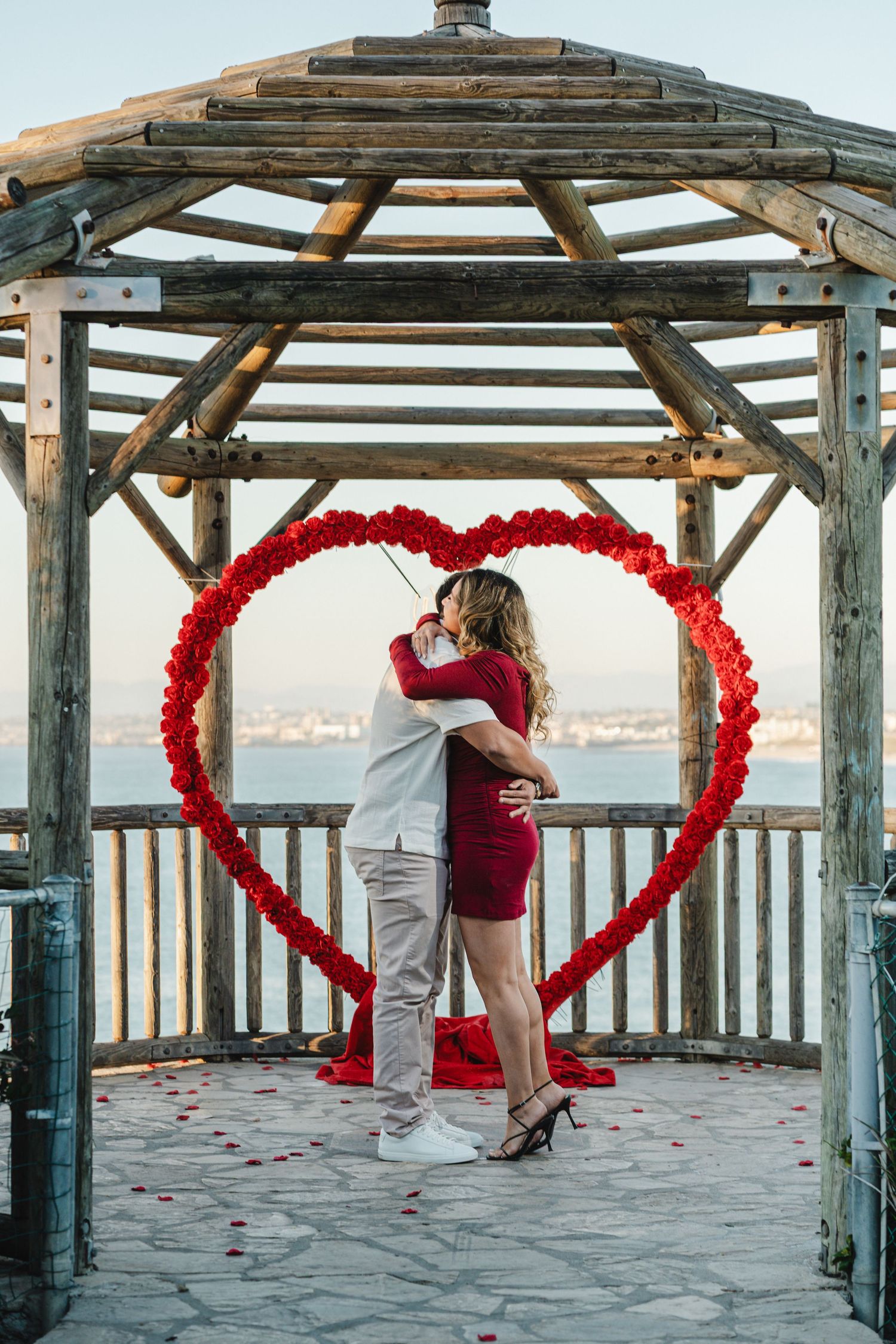 Romantic proposal setup with red heart arch and rose petals on wooden pier overlooking ocean.
