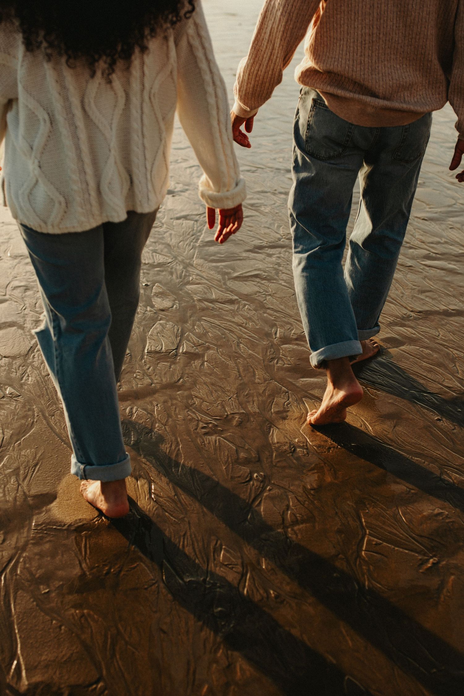 Multiple people walking barefoot on wet sand at the beach during sunset, casting long shadows on the shoreline.
