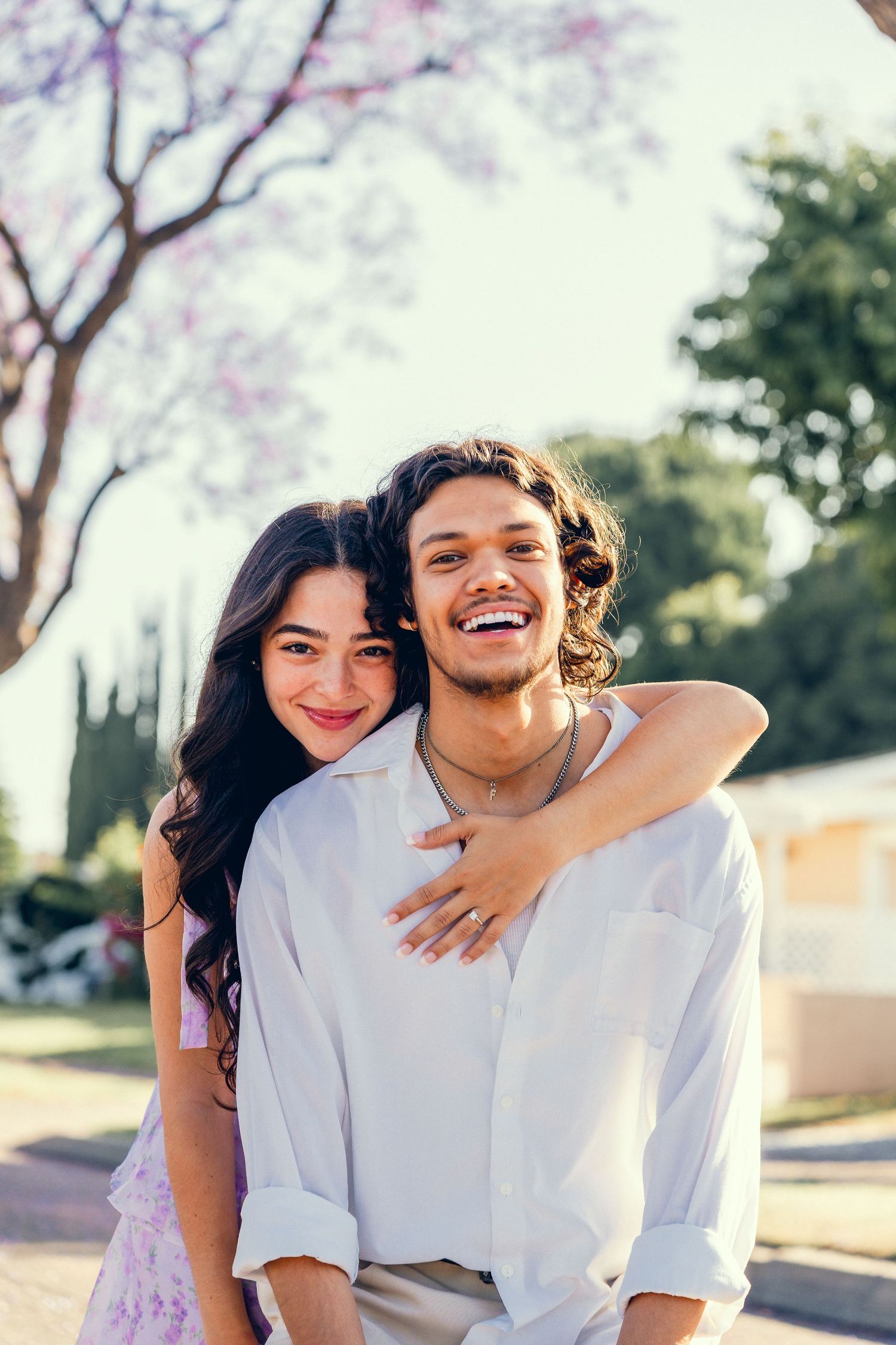 Couple in white outfits embraces under blooming purple jacaranda trees.