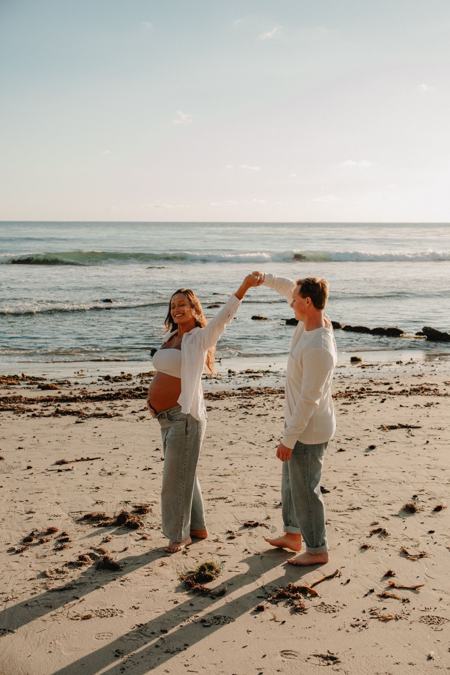 A couple shares a tender moment while walking along a sandy beach at sunset with ocean waves in the background.