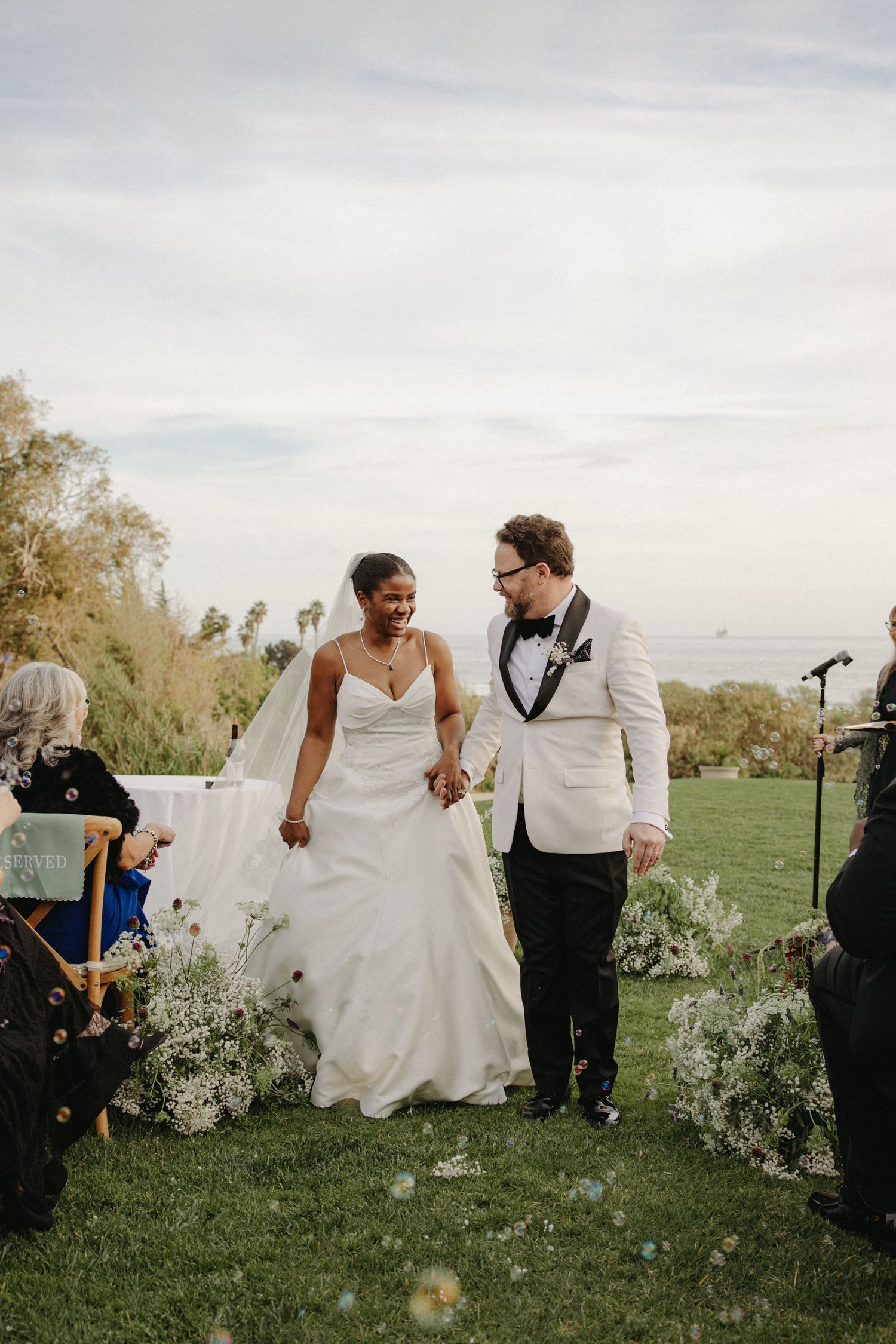 Bride and groom walk down the aisle hand in hand at an outdoor wedding ceremony with ocean views.
