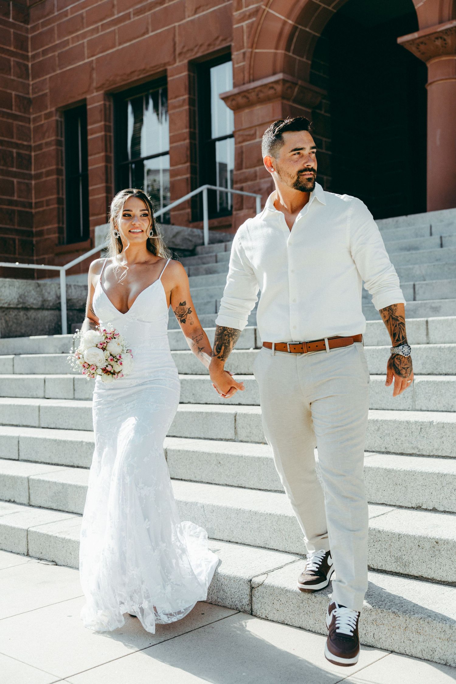 A couple descends white stone steps outside a brick building in matching white wedding attire.