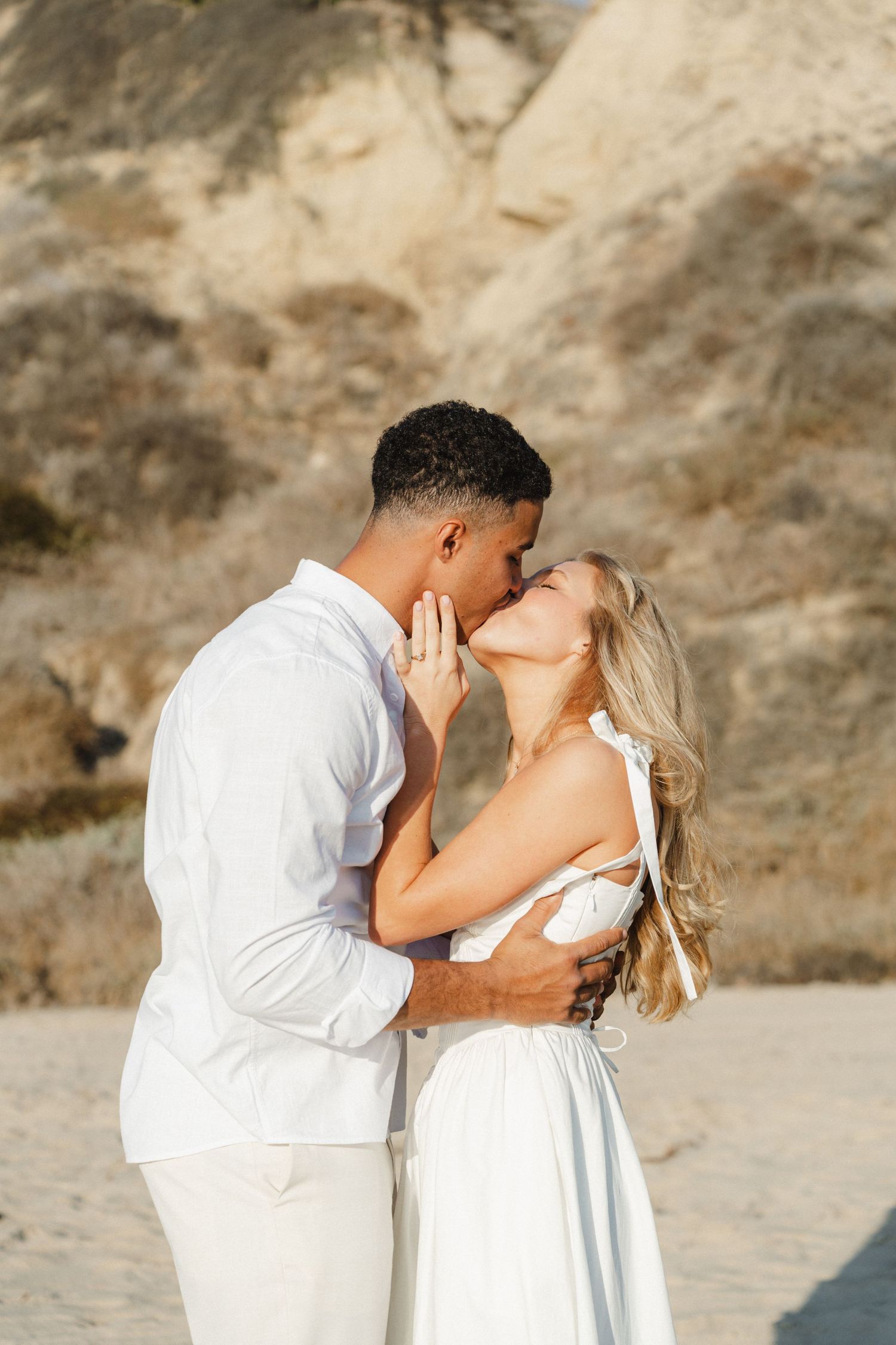 Romantic beach portraits of couple in white attire against rocky coastal backdrop.