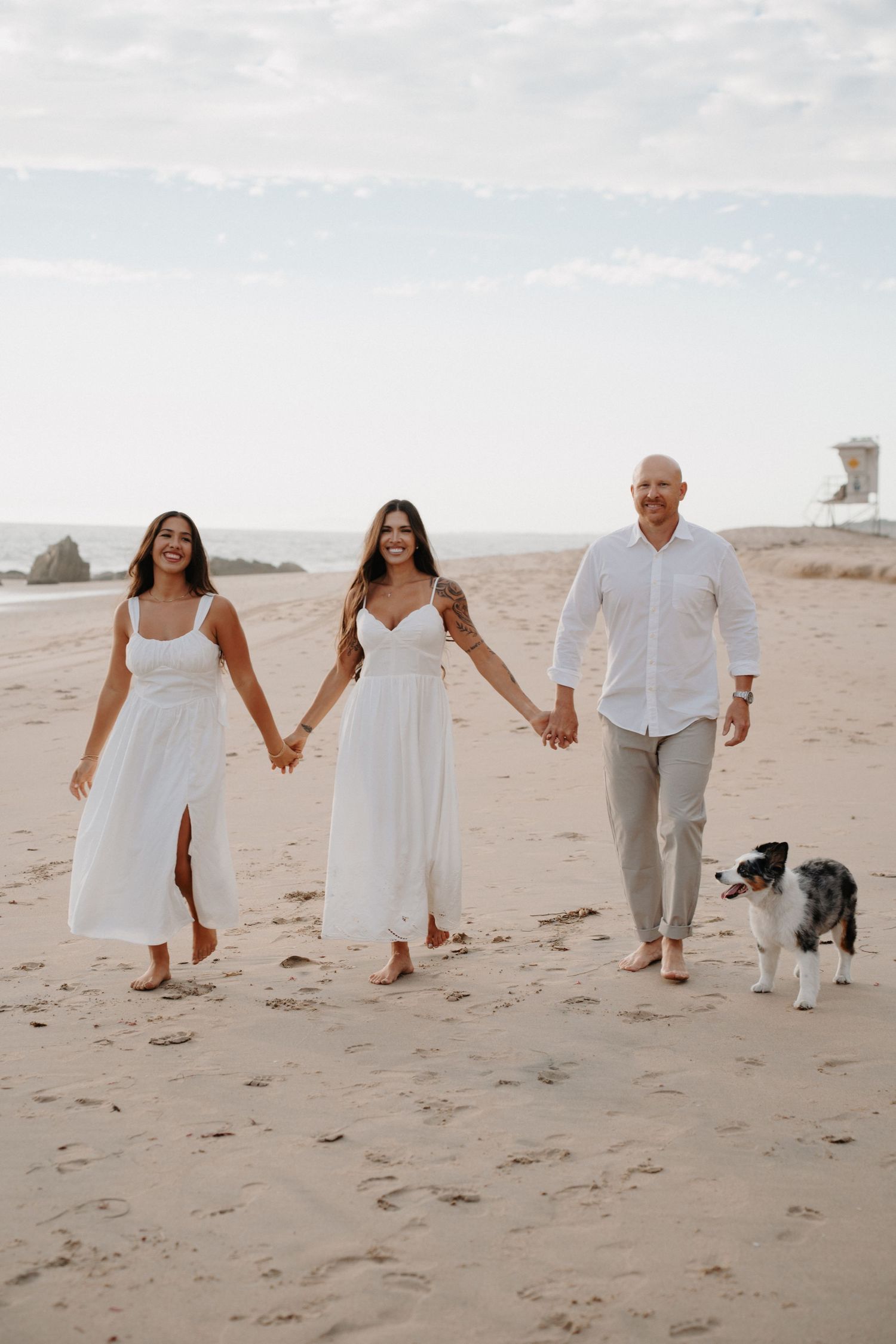 Three people in white outfits walk together with a border collie dog on a sandy beach at sunset.