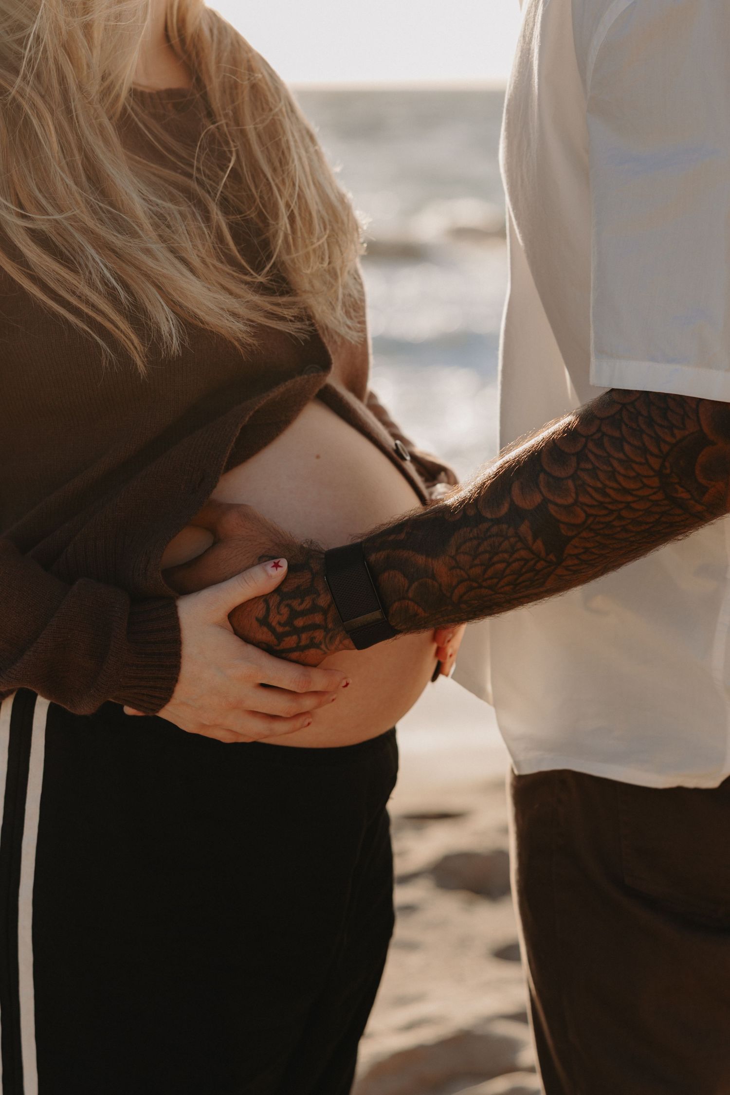 Close-up view of hands tenderly cradling a pregnant belly on a beach at sunset.