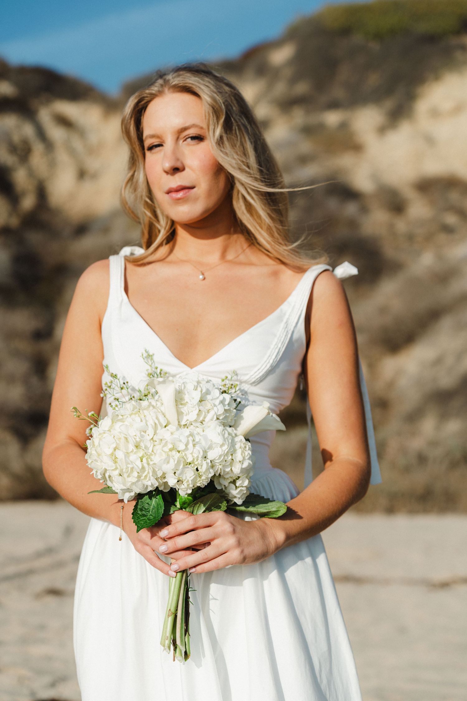 Bride in white dress holds bouquet of white flowers against coastal cliff backdrop.