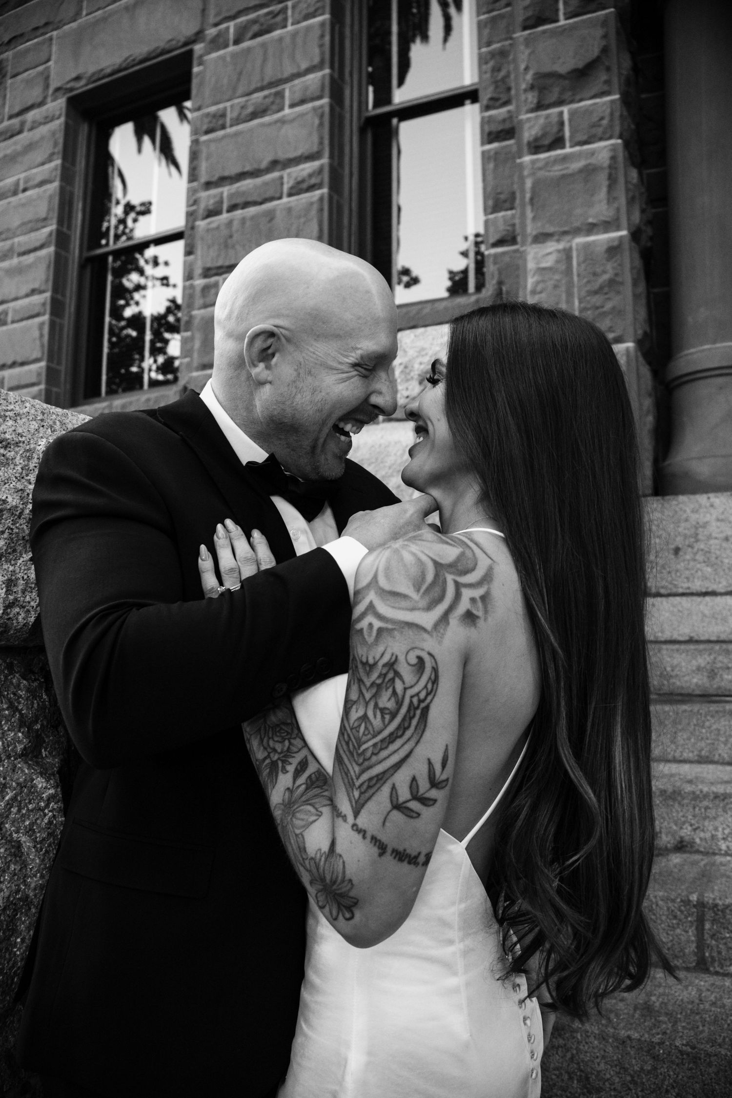 Black and white wedding photo showing a joyful moment between a couple, with the bride's back tattoo visible in her dress.