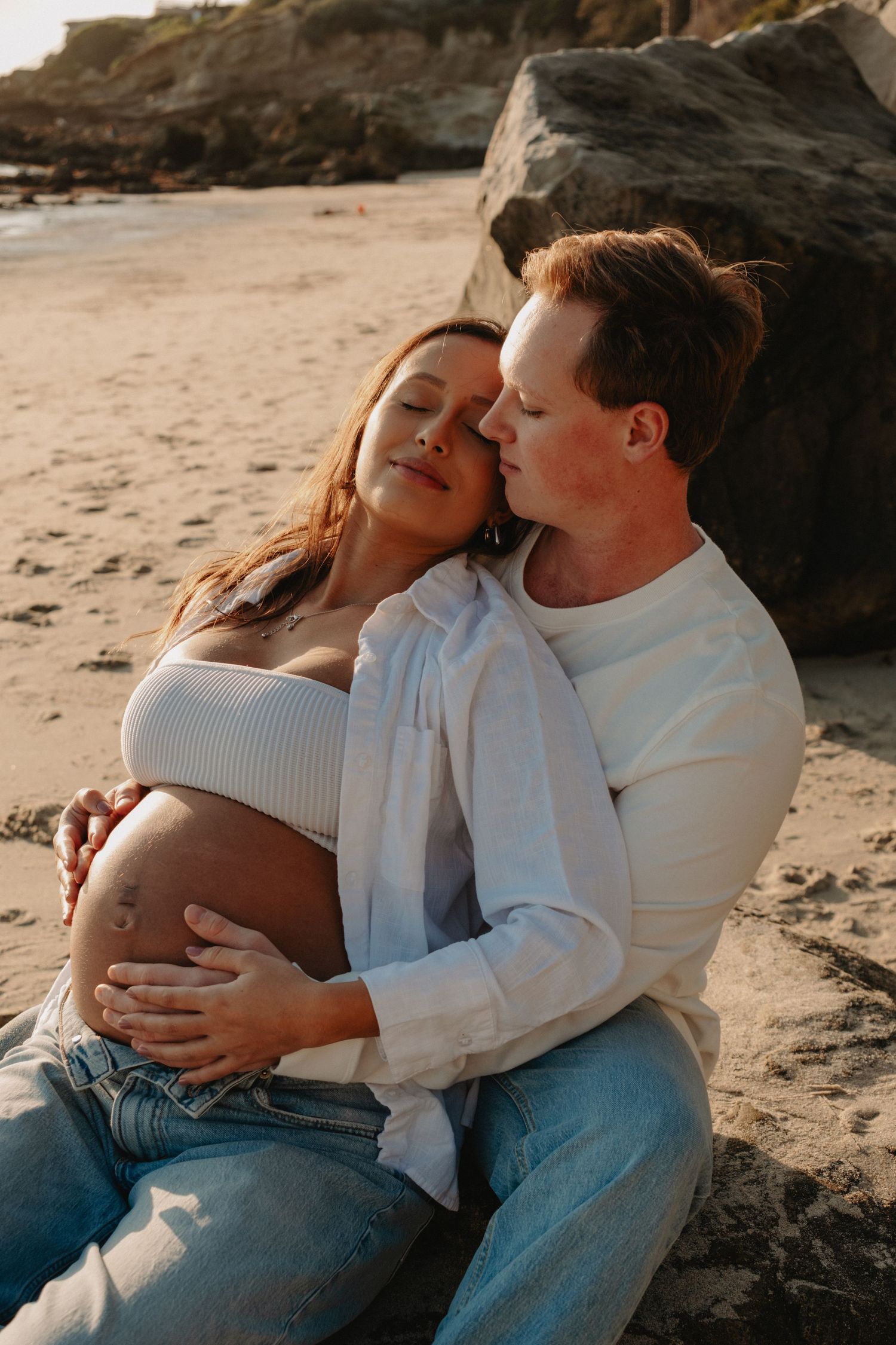 A couple in white sweaters and jeans share intimate moments on a sandy beach at sunset.
