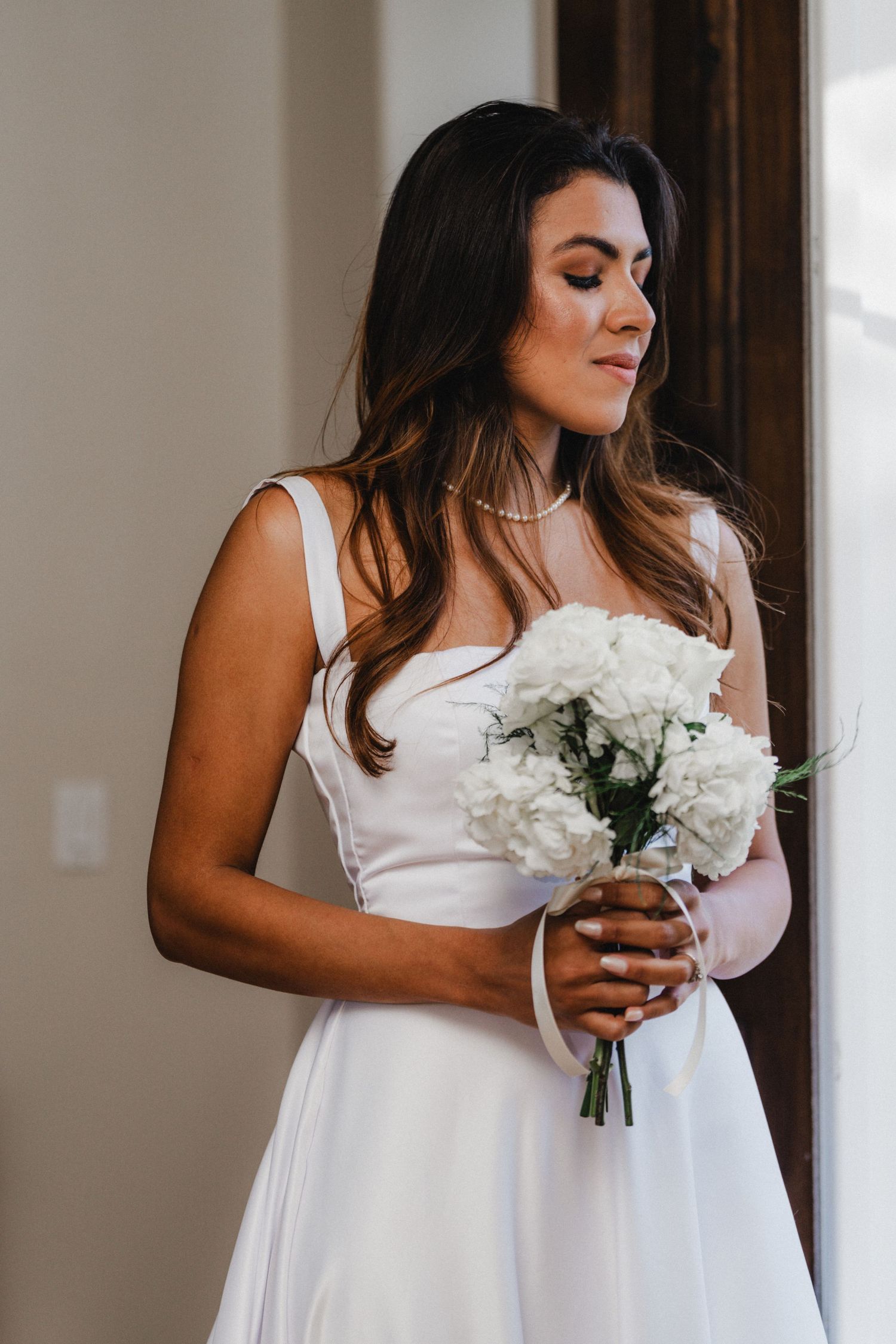 Person in white wedding dress holding elegant white floral bouquet in indoor setting.