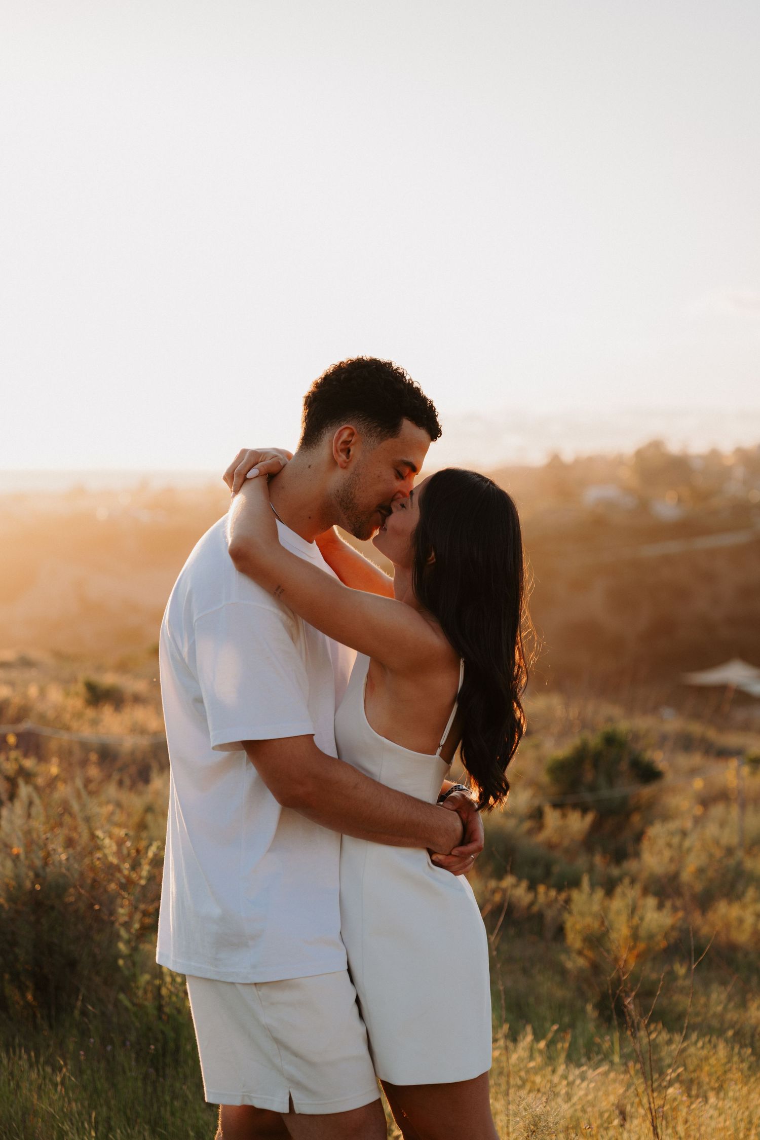 Couple in white clothing embracing during sunset in a grassy field.