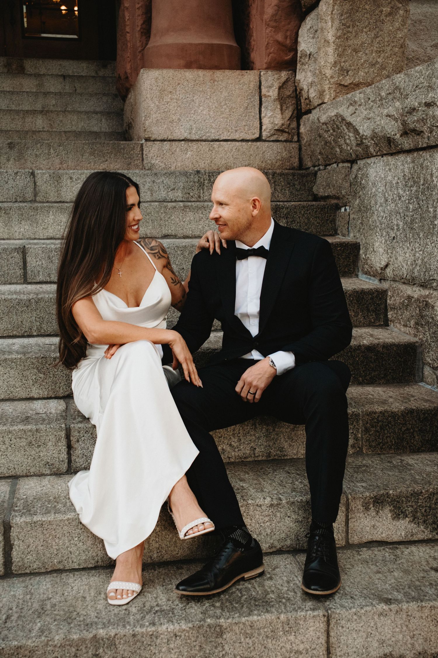 A couple in wedding attire sits together on stone steps sharing an intimate moment in a romantic outdoor setting.