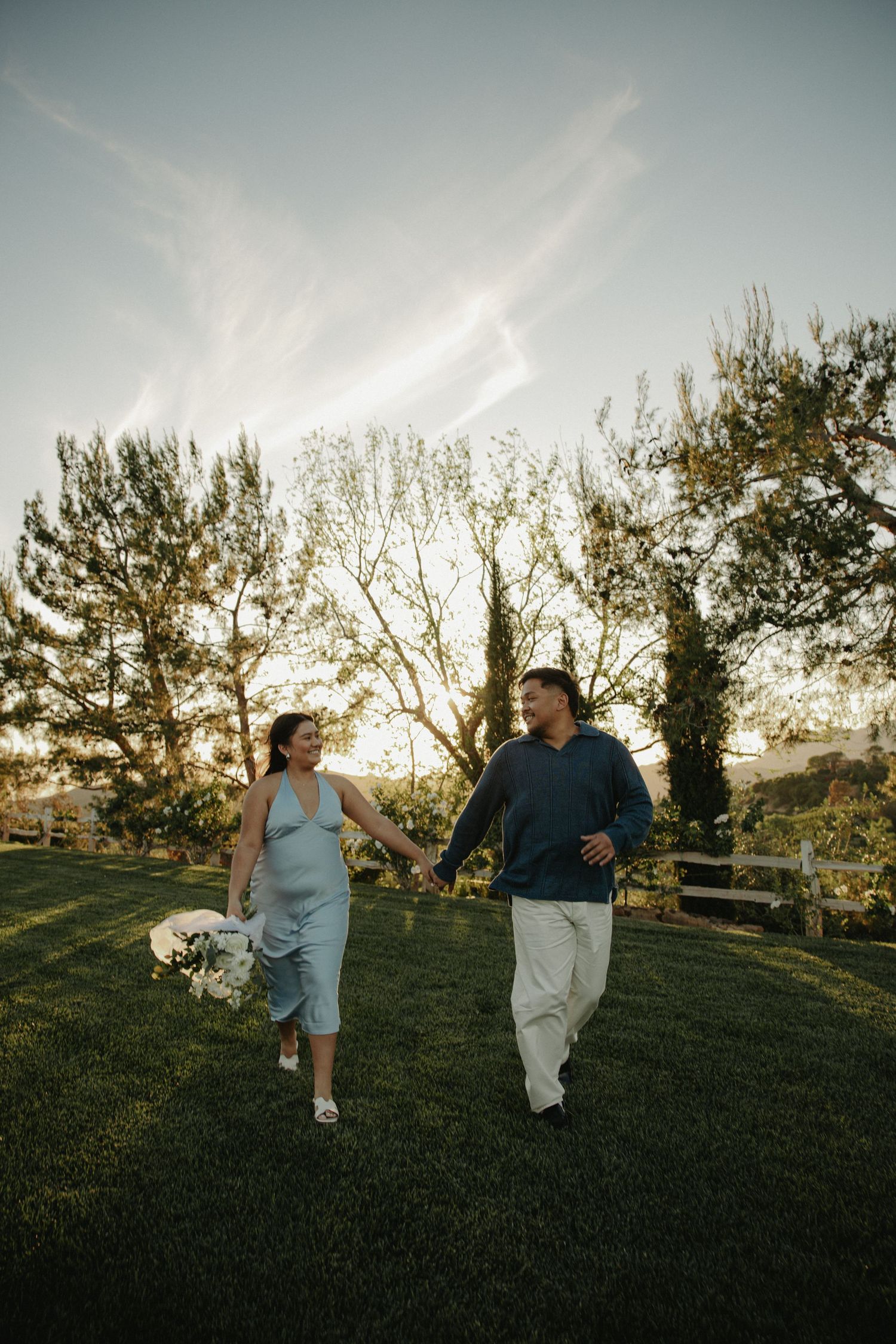 A couple walks hand-in-hand on a grassy lawn at sunset, woman in blue dress holding flowers, man in navy shirt.
