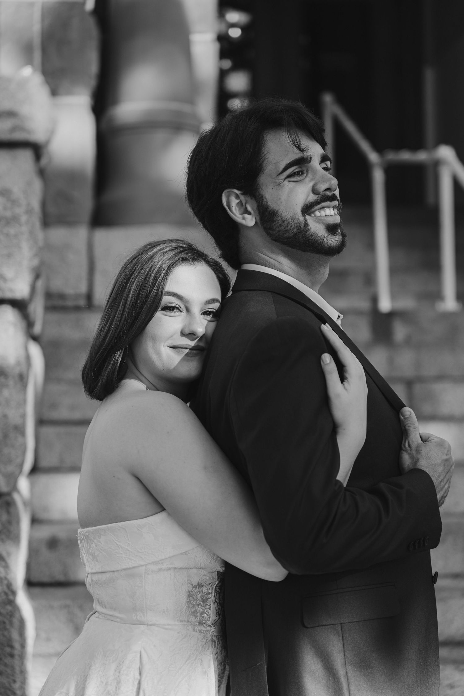 A couple shares joyful embraces and laughter in front of a stone building.
