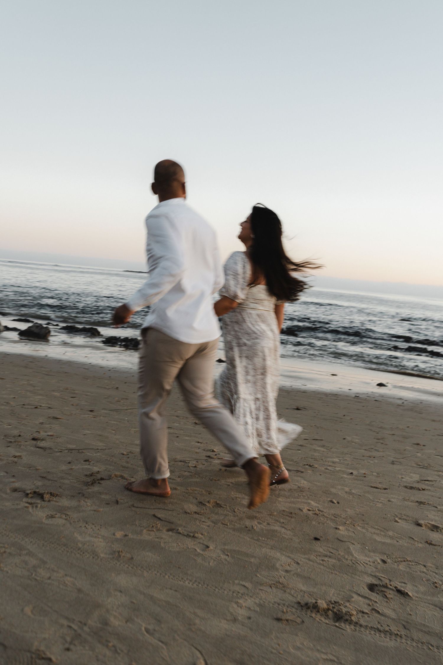 A couple in white clothing runs joyfully along a beach at sunset.