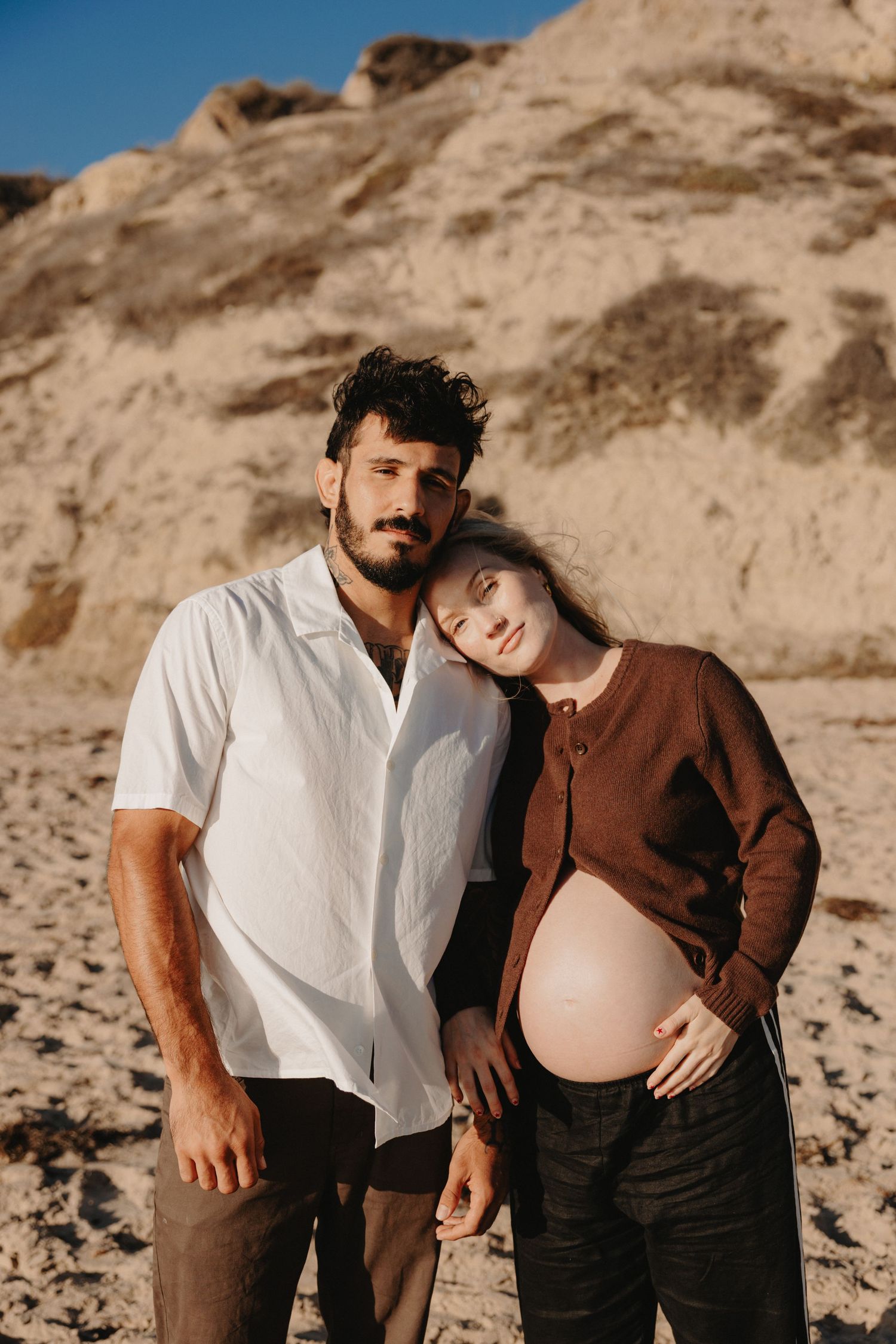 A couple in casual attire poses together on a sandy beach during a maternity photoshoot at sunset.