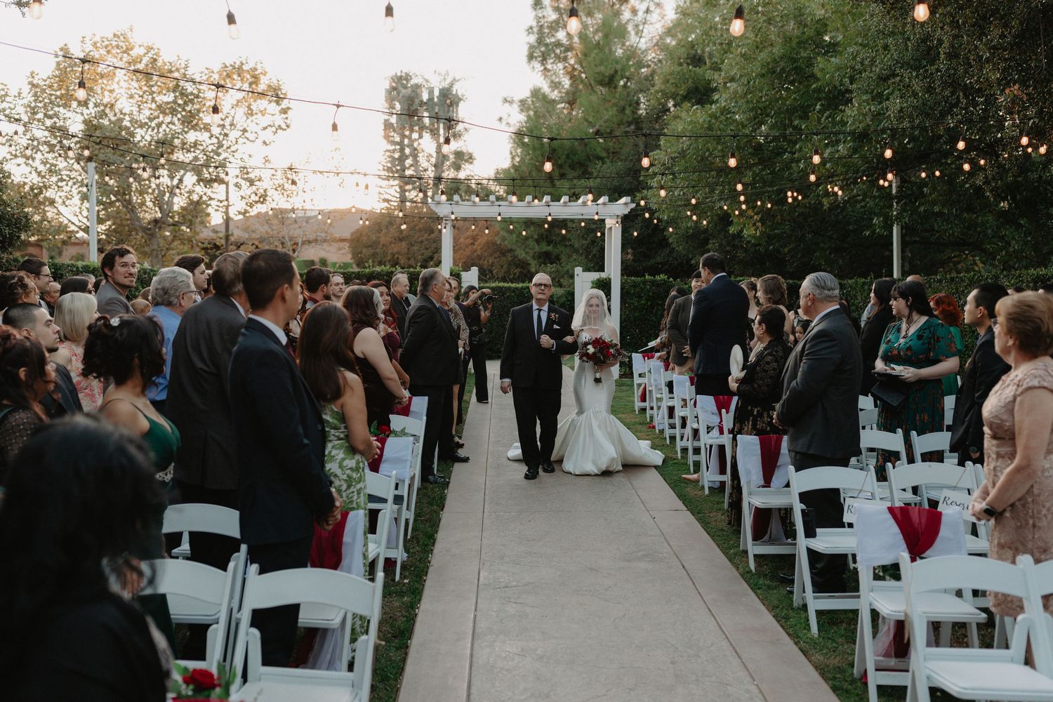 Outdoor evening wedding ceremony under string lights with guests seated in white chairs along a concrete pathway.
