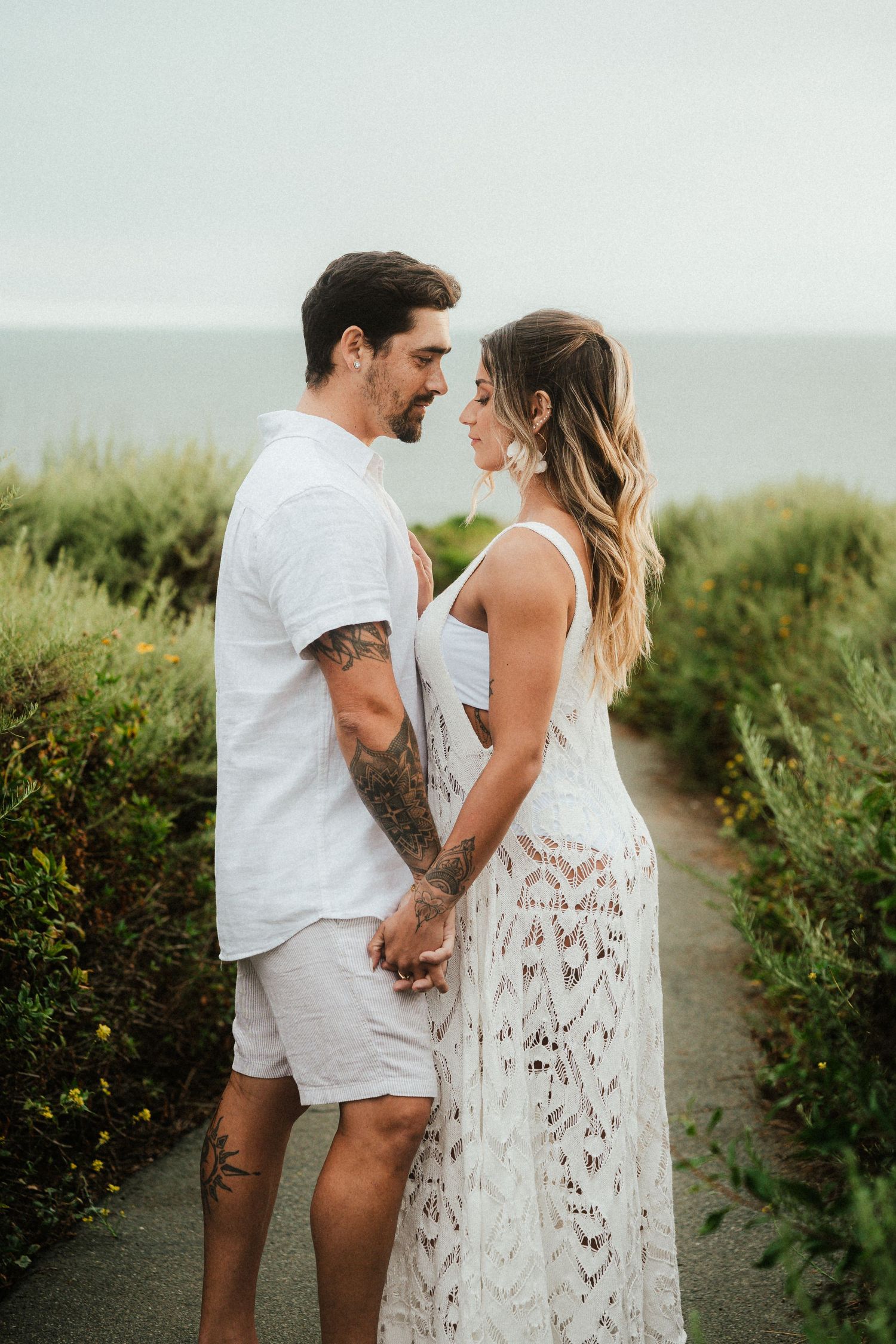 A couple in white outfits shares an intimate moment on a coastal path with ocean views.