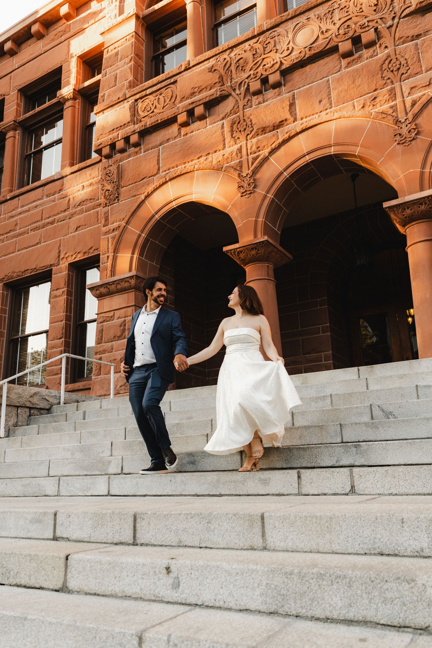 Couple in formal attire playfully interacting on grand stone steps of historic building.