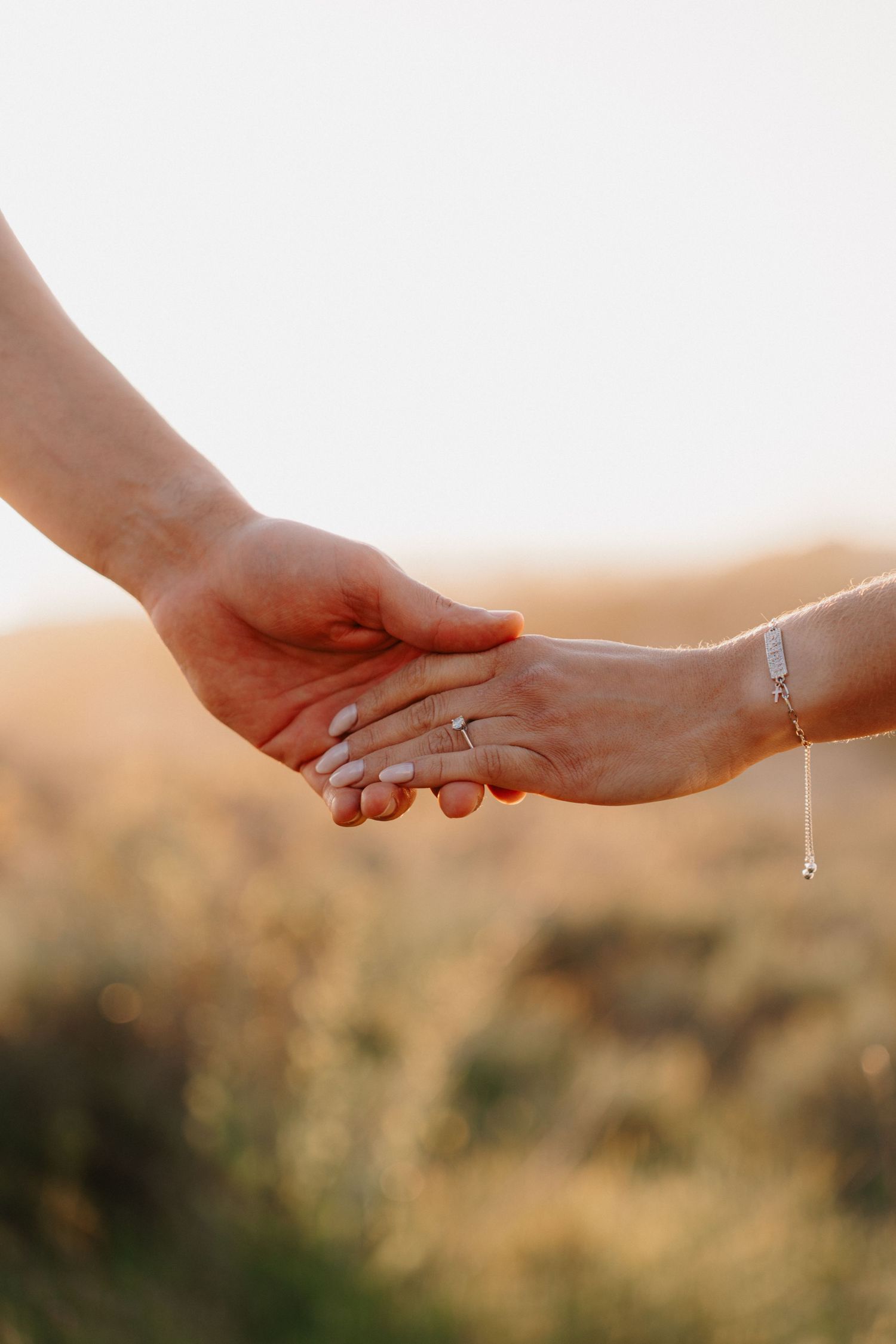 Two hands reaching out to each other against a blurred golden sunset background, one wearing a delicate bracelet.