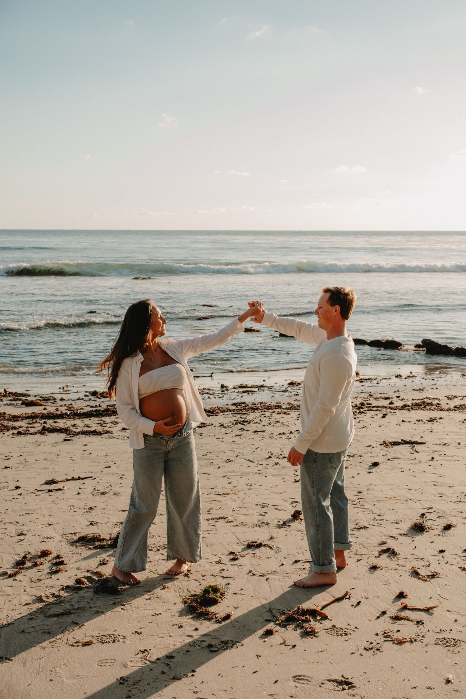 Two people standing on a beach at sunset holding hands while wearing casual white and gray outfits.