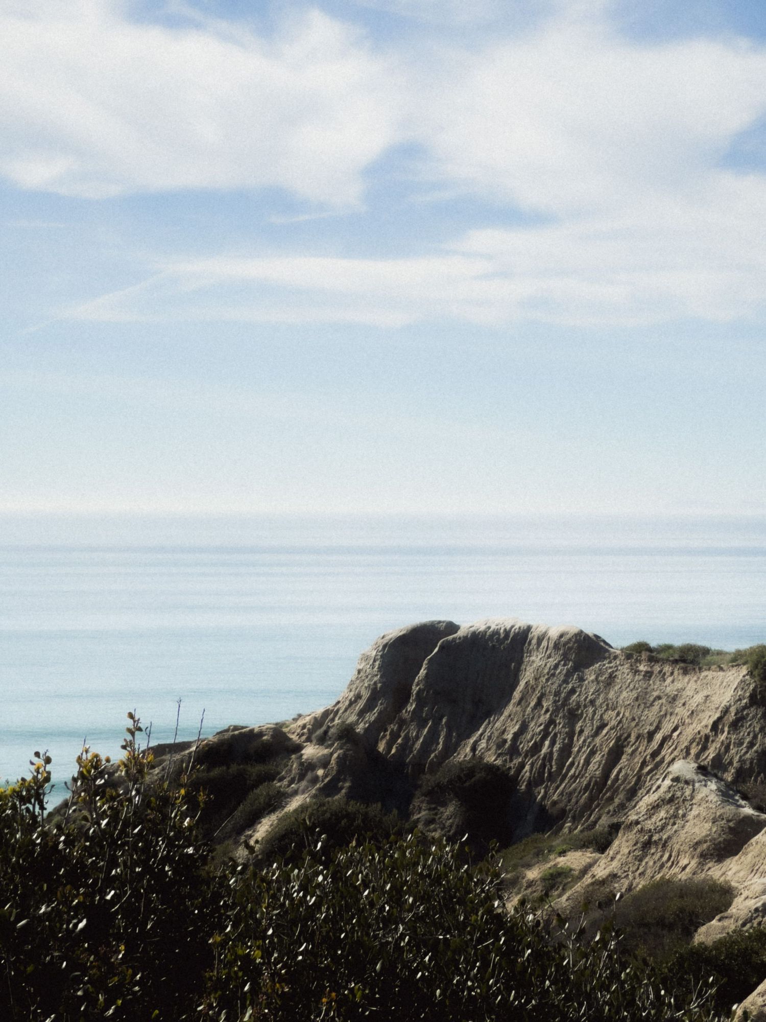 Coastal cliff overlooking calm blue ocean under wispy clouds.