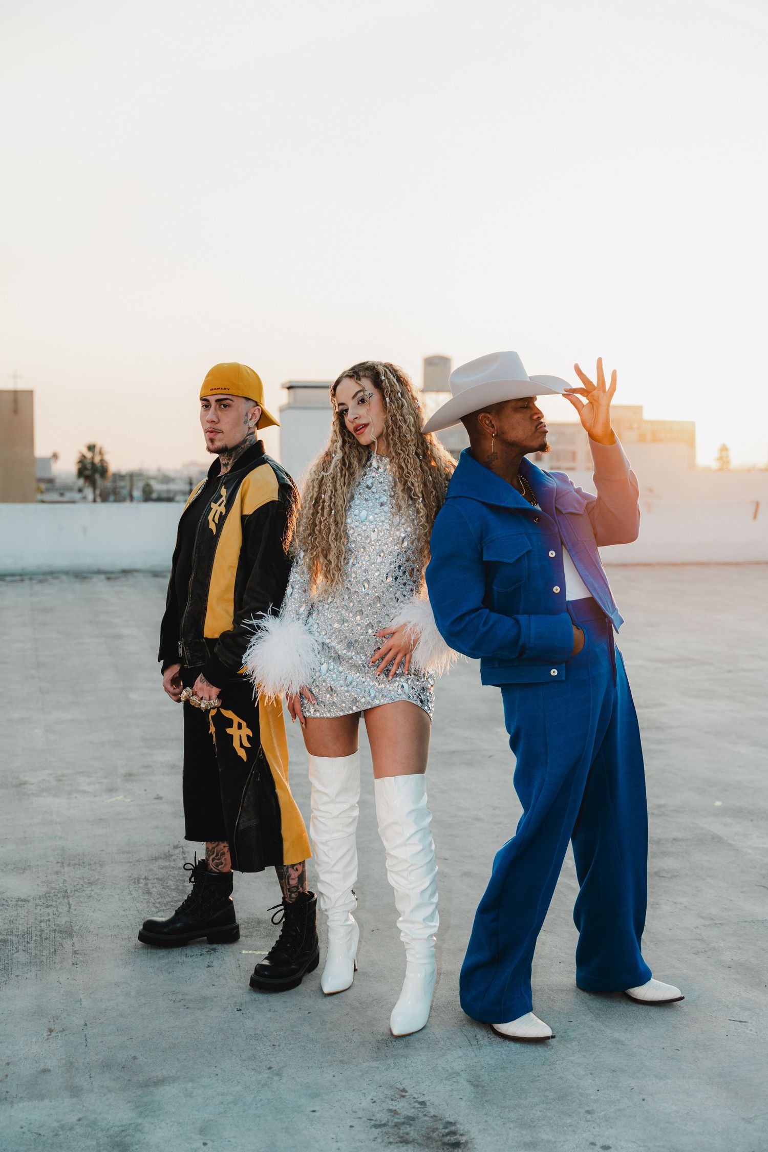 Three stylish performers pose on rooftop in contrasting outfits during golden hour.