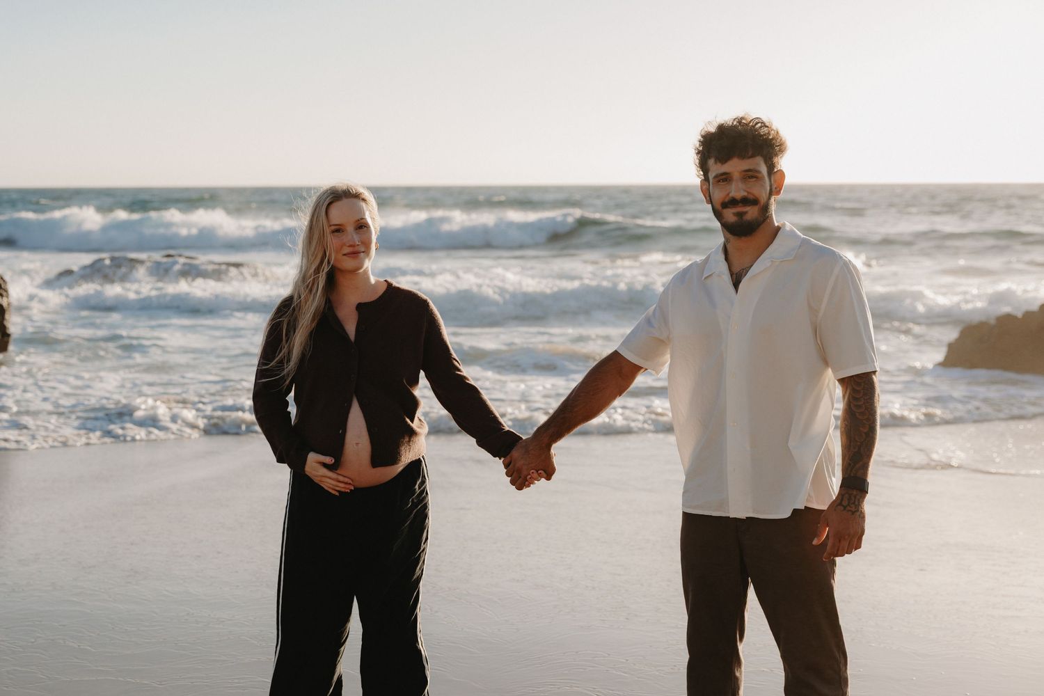 Couple walking together on a scenic beach with ocean waves crashing in the background during sunset.