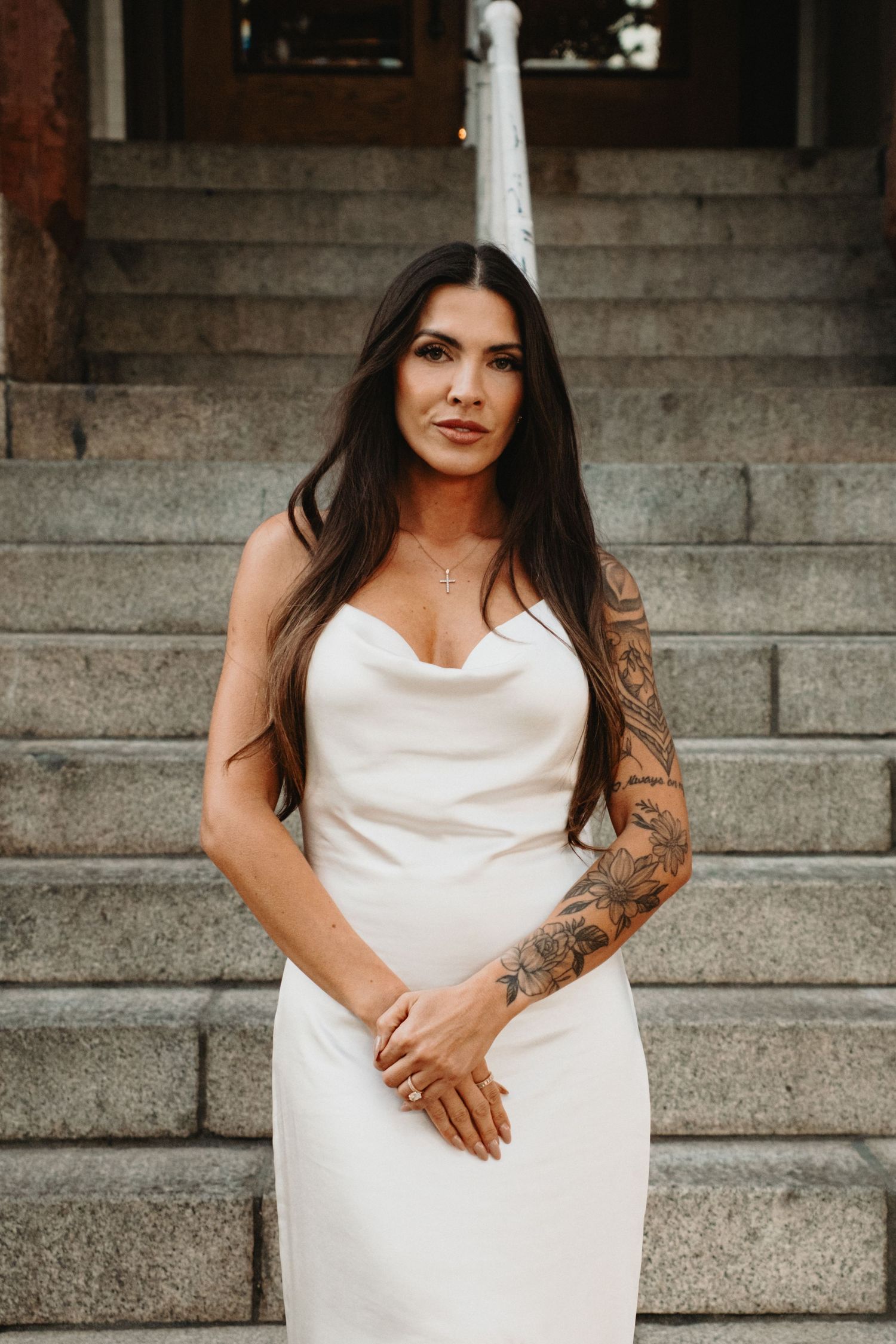 A person in a white satin dress poses elegantly on stone steps, showing detailed arm tattoos.