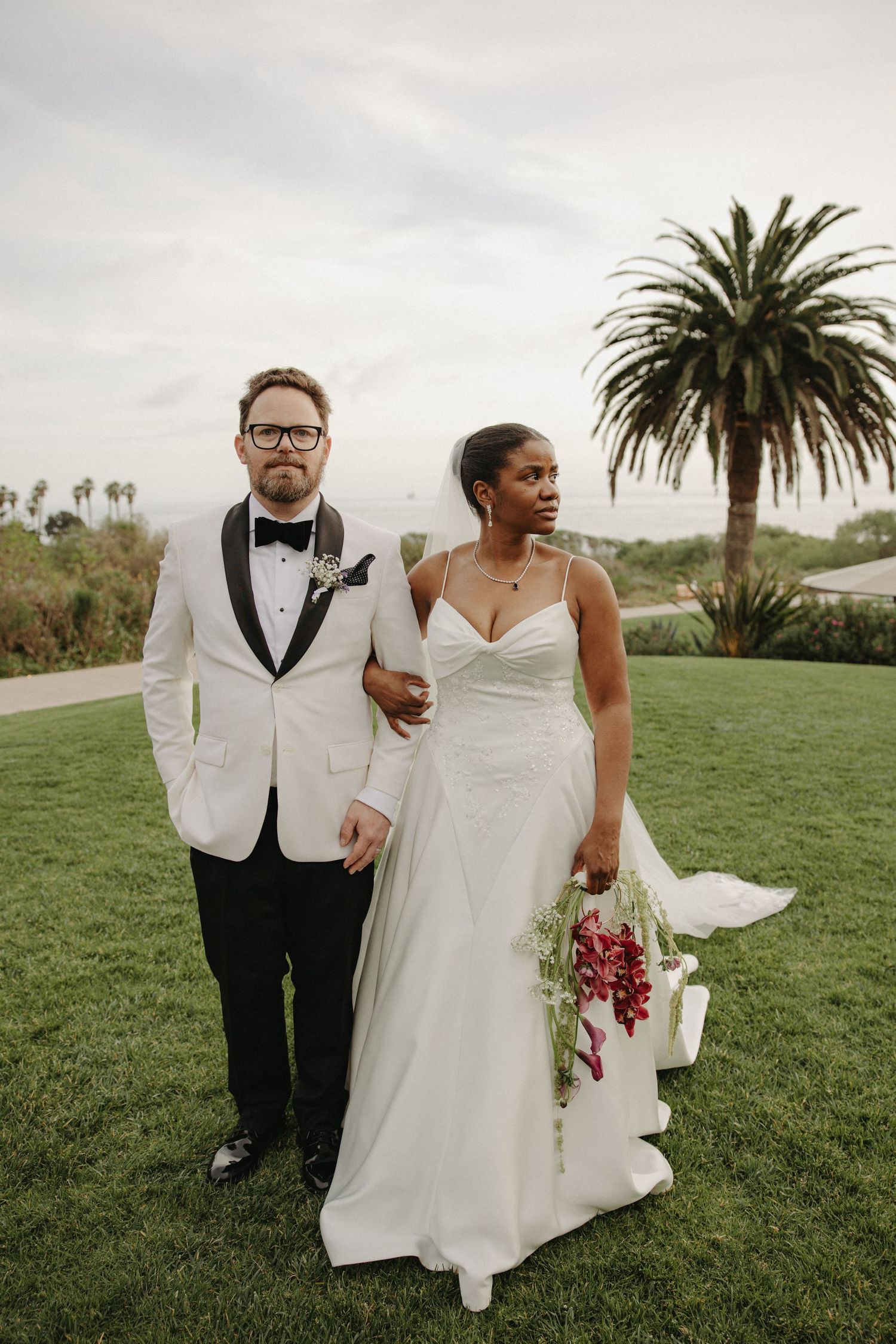 Bride and groom pose on green lawn, groom in white tuxedo jacket, bride in elegant white gown holding red bouquet.
