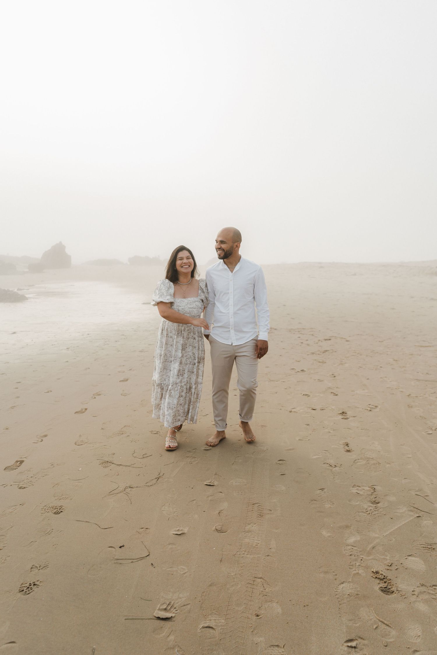 A couple takes a romantic walk along a foggy beach wearing matching light-colored outfits.