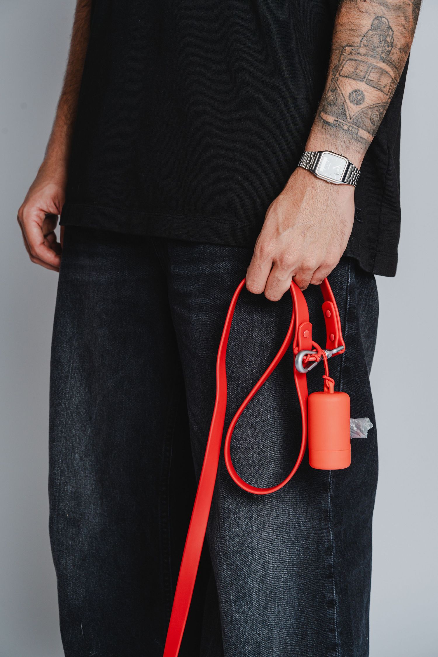 Person with tattoos holding a bright red dog leash against grey background.